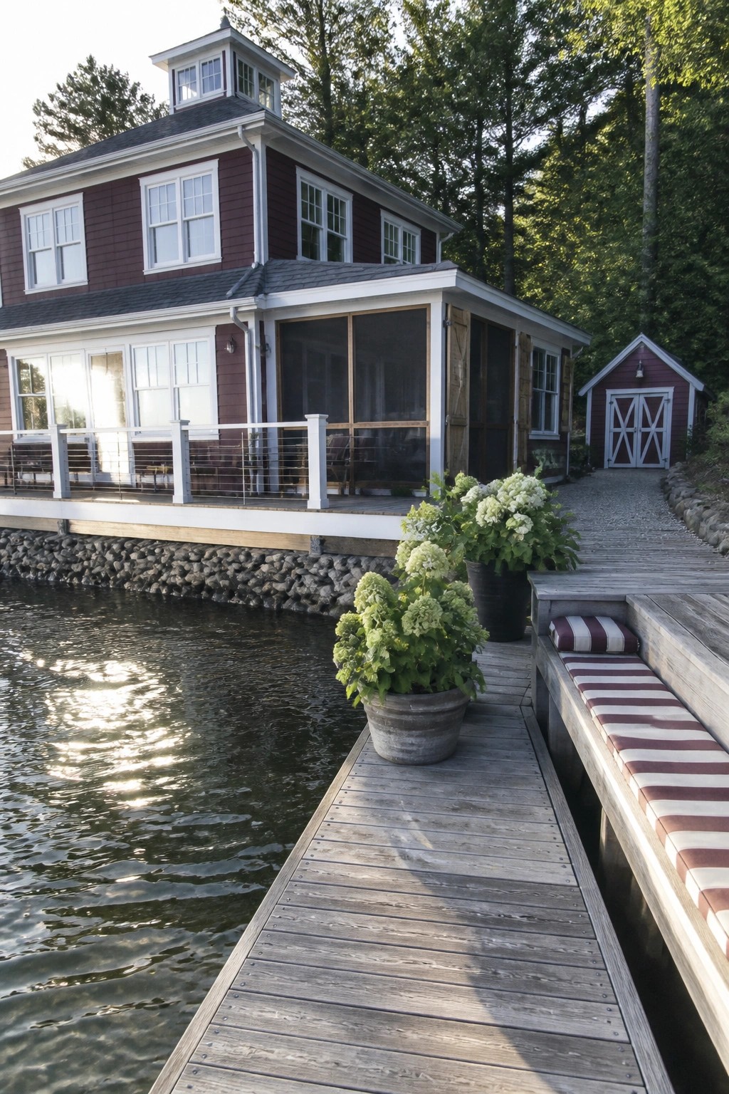 A two-story house with deep red lap siding and white trim stands beside a wooden dock, featuring a screened porch and potted plants along the water's edge.