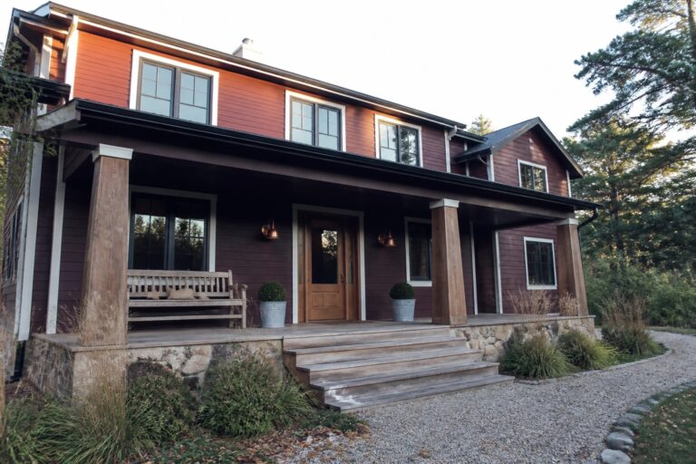21 Striking Maroon House Exteriors Using Deep Red Lap Siding A two-story house with deep red horizontal siding, a wooden front door, and a front porch supported by thick wooden posts.