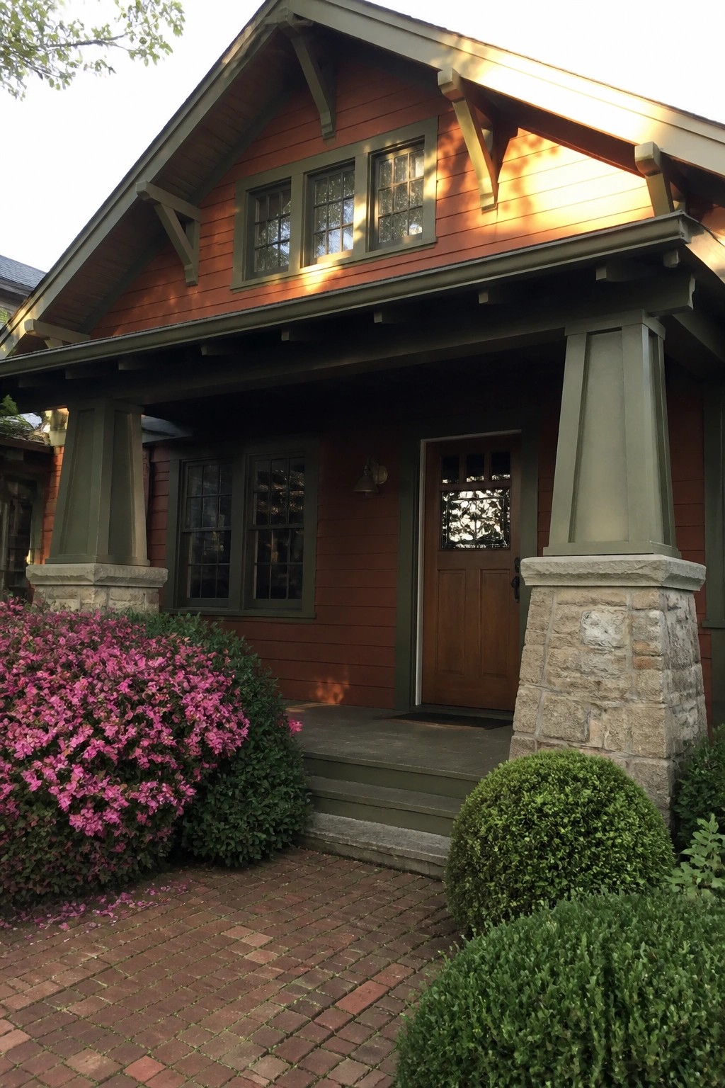 Red wooden house with green trim, stone pillars, pink azaleas, and brick path.