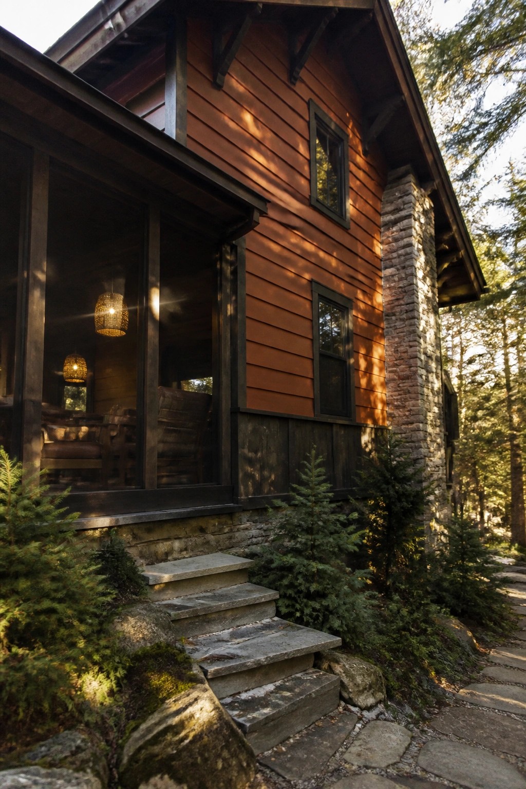 A house exterior featuring burnt orange lap siding, dark window trim, a stone chimney, and stone steps leading to a covered porch area.