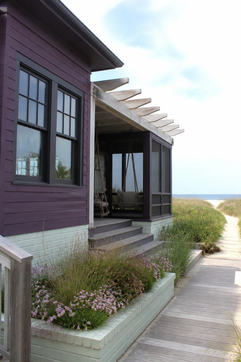 A house exterior with deep purple siding, dark-framed windows, a screened porch, and steps leading to a wooden walkway near the beach.