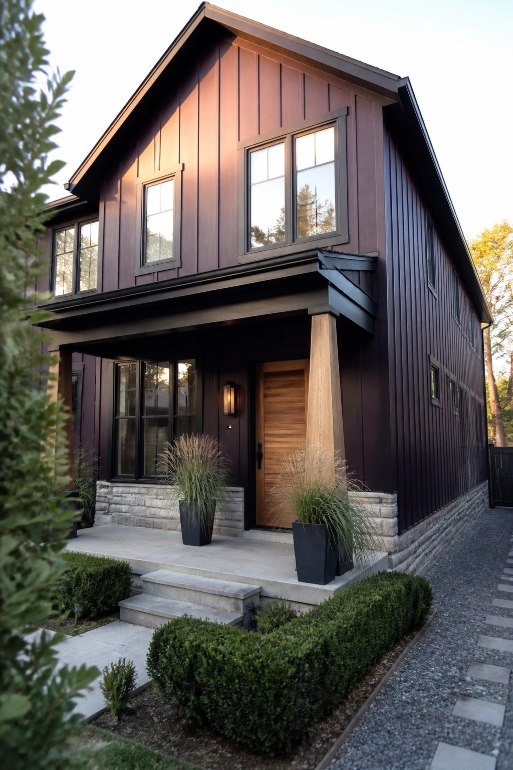 A two-story modern house with aubergine vertical siding, black trim, a wooden front door, stone foundation, and two large potted grasses on the concrete porch.