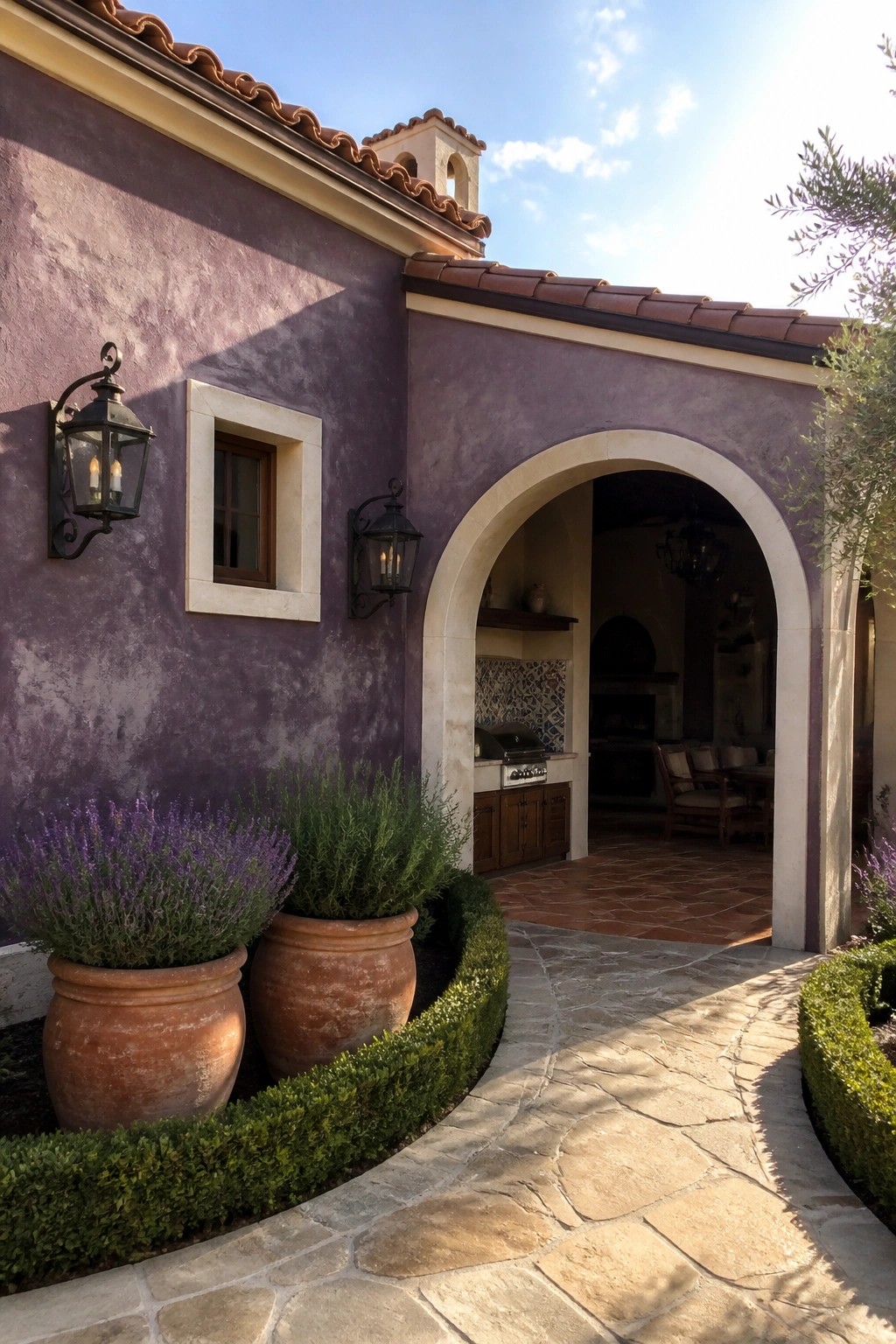 A purple stucco house exterior with an arched entryway, terracotta roof tiles, terracotta pots holding plants, and a curved stone pathway.
