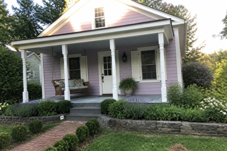 Front view of a light purple house with white trim, porch columns, white door, and stone steps, with landscaping in the foreground.