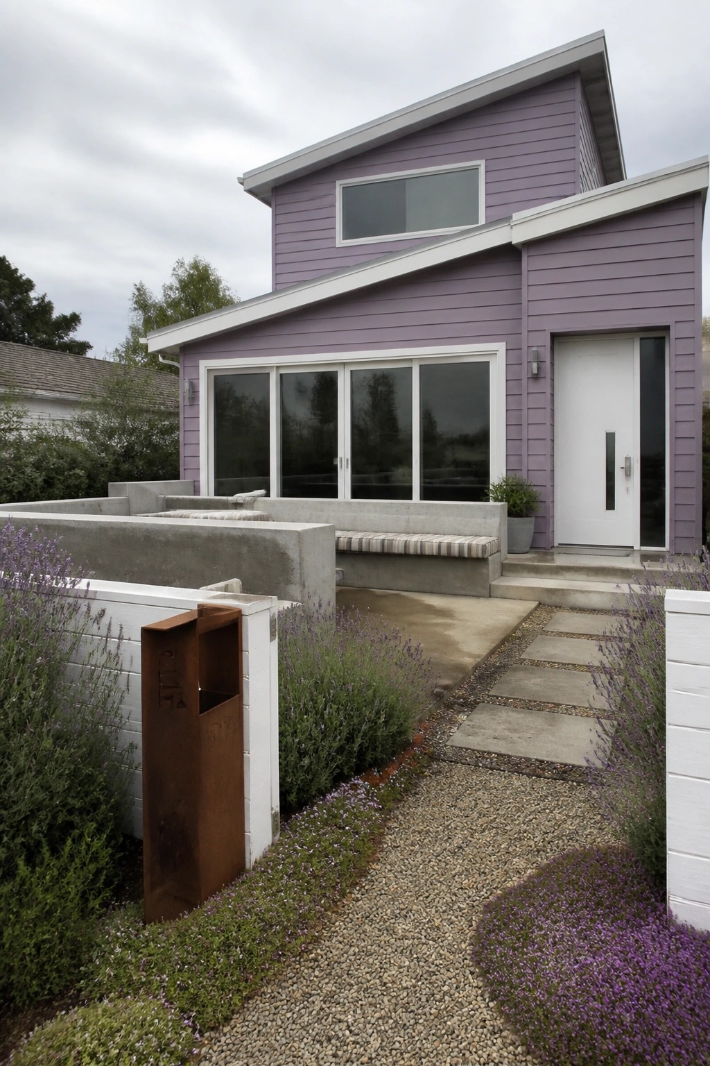 A two-story house with lilac siding, white trim around windows and edges, a white front door, concrete steps, and a gravel path bordered by lavender plants.