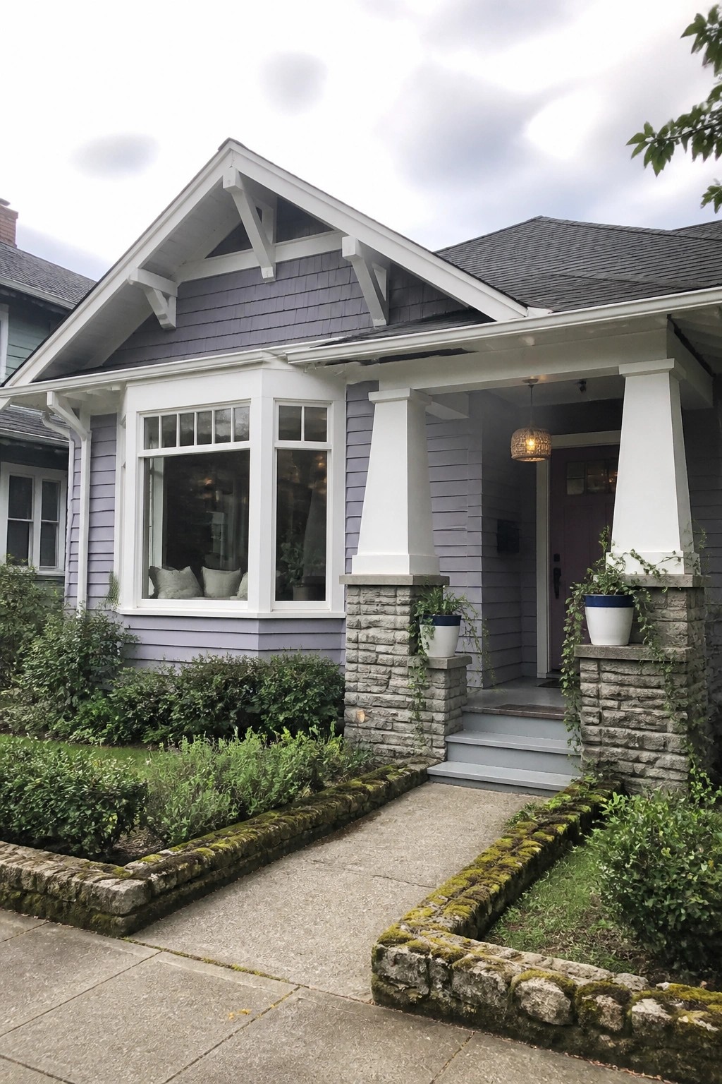 Front exterior of a lilac house with white trim, white porch columns on stone bases, and a concrete walkway bordered by low stone edging.