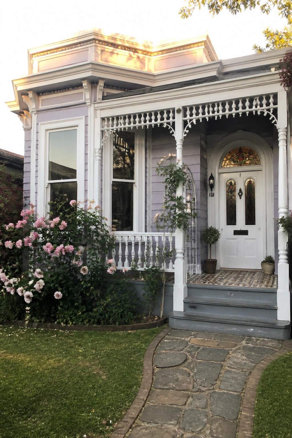 A lilac house exterior with white trim shows a front porch, white door with stained glass panels, and climbing roses along the side.