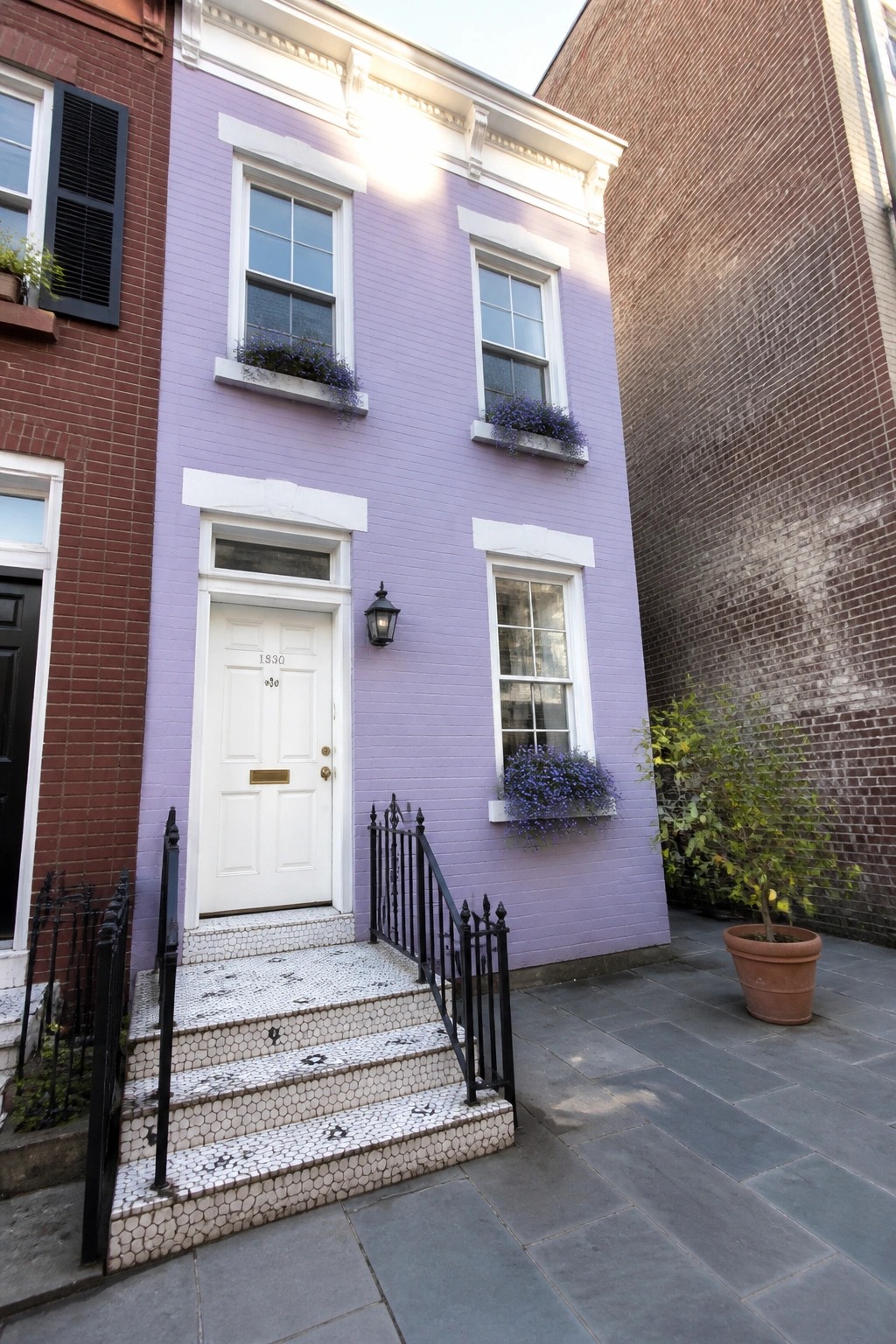 A narrow lilac brick house with white trim on windows and door, white paneled door, mosaic tile steps, and black railings.