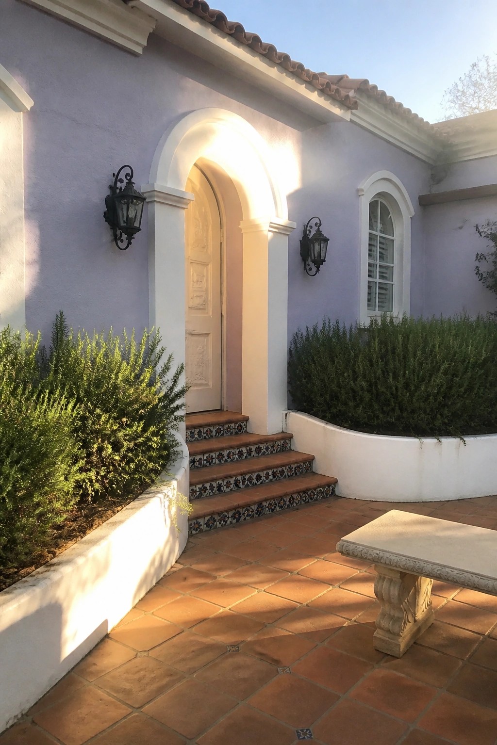 Purple stucco walls, arched doorway, terracotta tiles, stone bench in sunny courtyard.