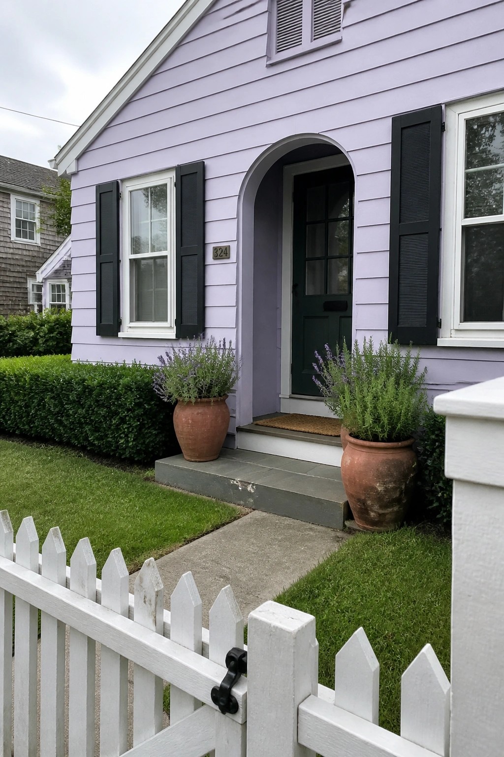 Lavender cottage exterior with black shutters, an arched entryway around a dark door, and two large terracotta pots beside the steps.