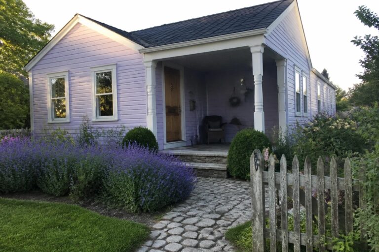 A single-story cottage with lavender siding, white trim, a wooden front door, stone steps, and a cobblestone walkway bordered by flowers and a wooden picket fence.