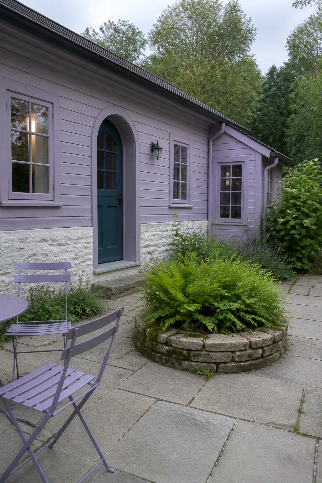 A lavender painted cottage with teal arched door, white stone foundation, stone patio, purple metal chairs, and a circular stone planter with ferns.