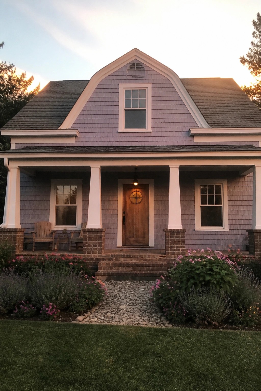 A lavender-shingled cottage with a white-trimmed front porch, brick steps, and a gravel walkway bordered by flower beds.