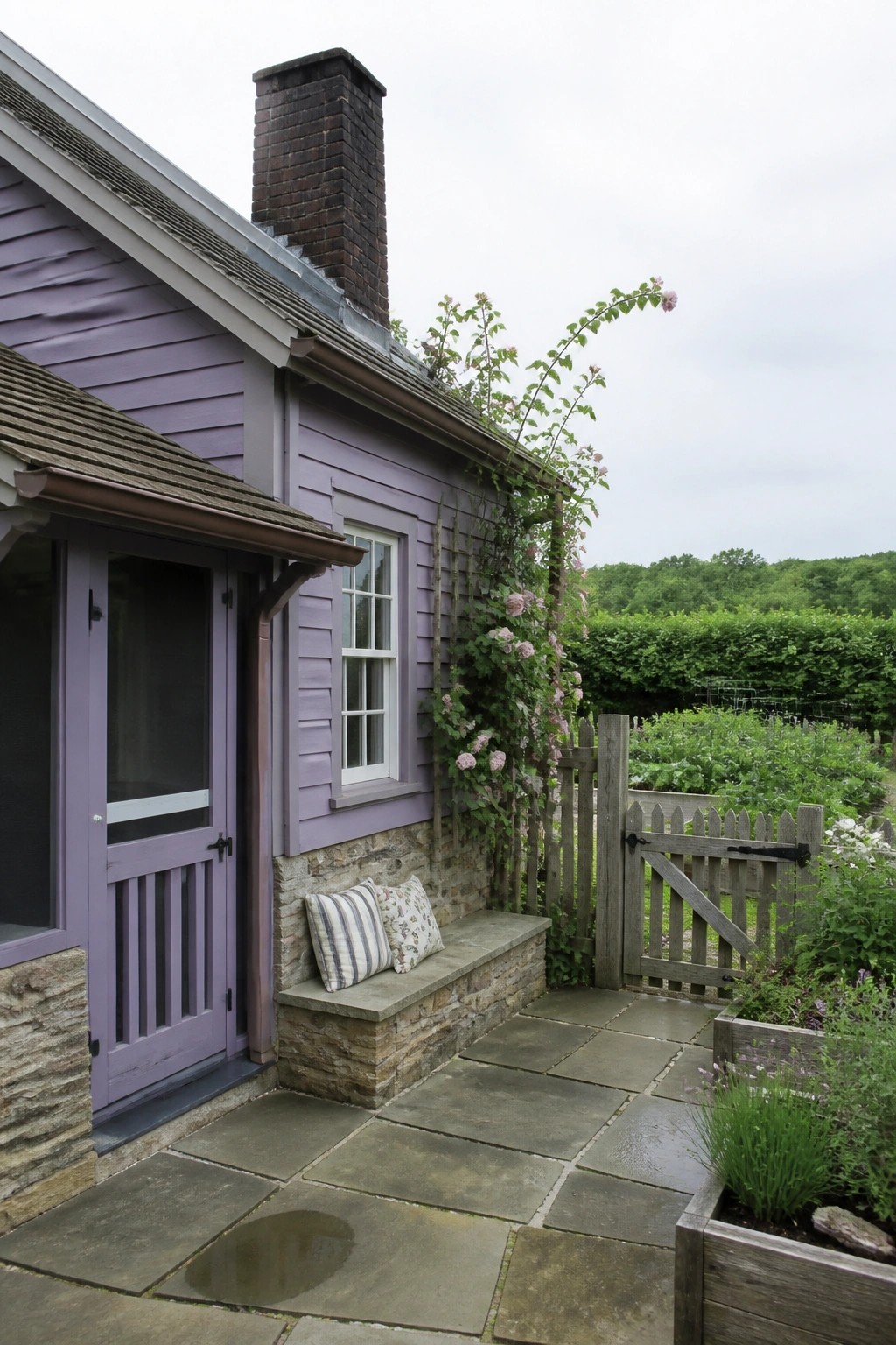 A lavender cottage exterior featuring a stone foundation, wooden door, window, chimney, stone bench with cushions, and a wooden gate opening to a garden.