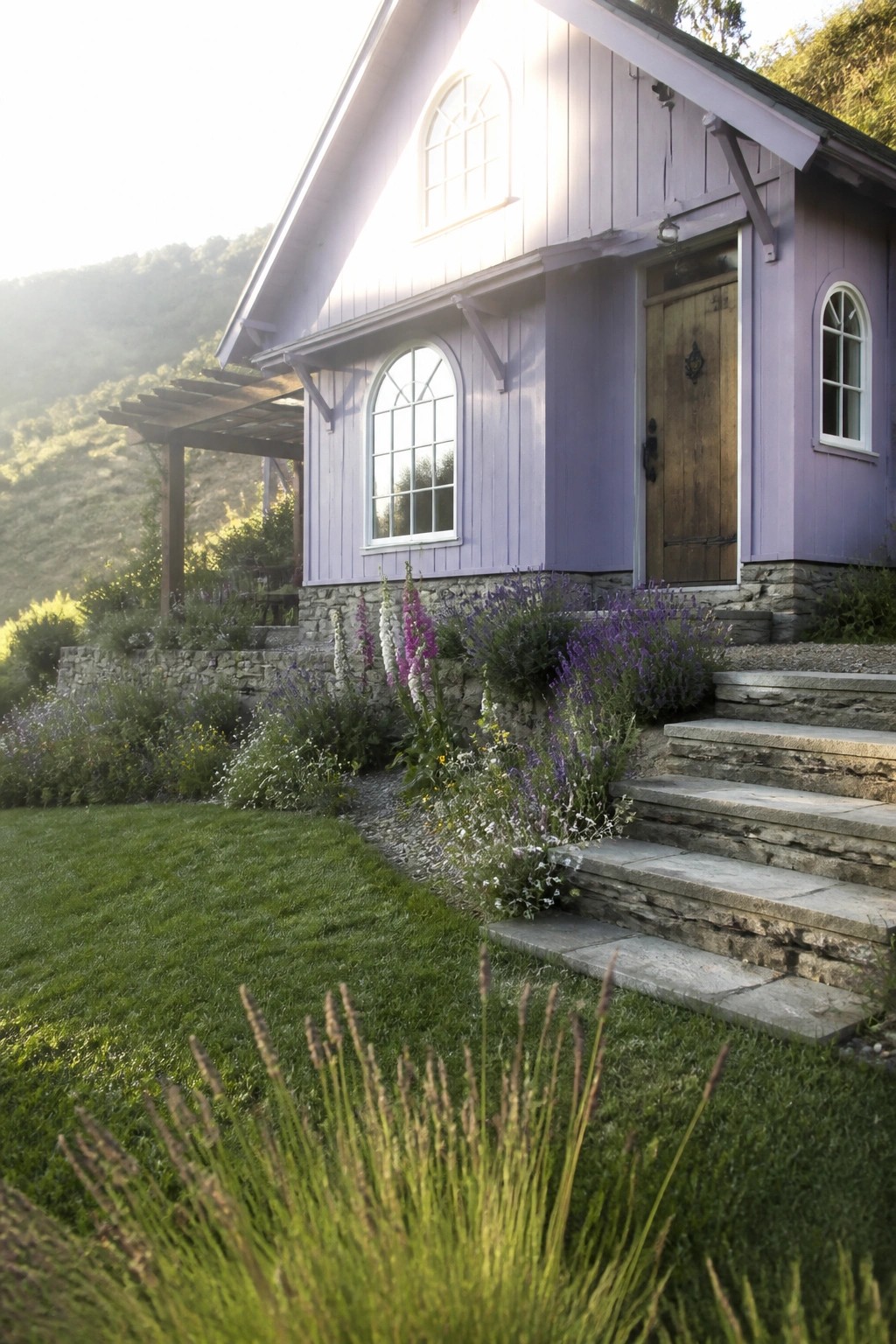 A lavender purple wooden cottage exterior with arched windows, a wooden front door, stone foundation, stone steps, and surrounding garden beds with purple and white flowers.