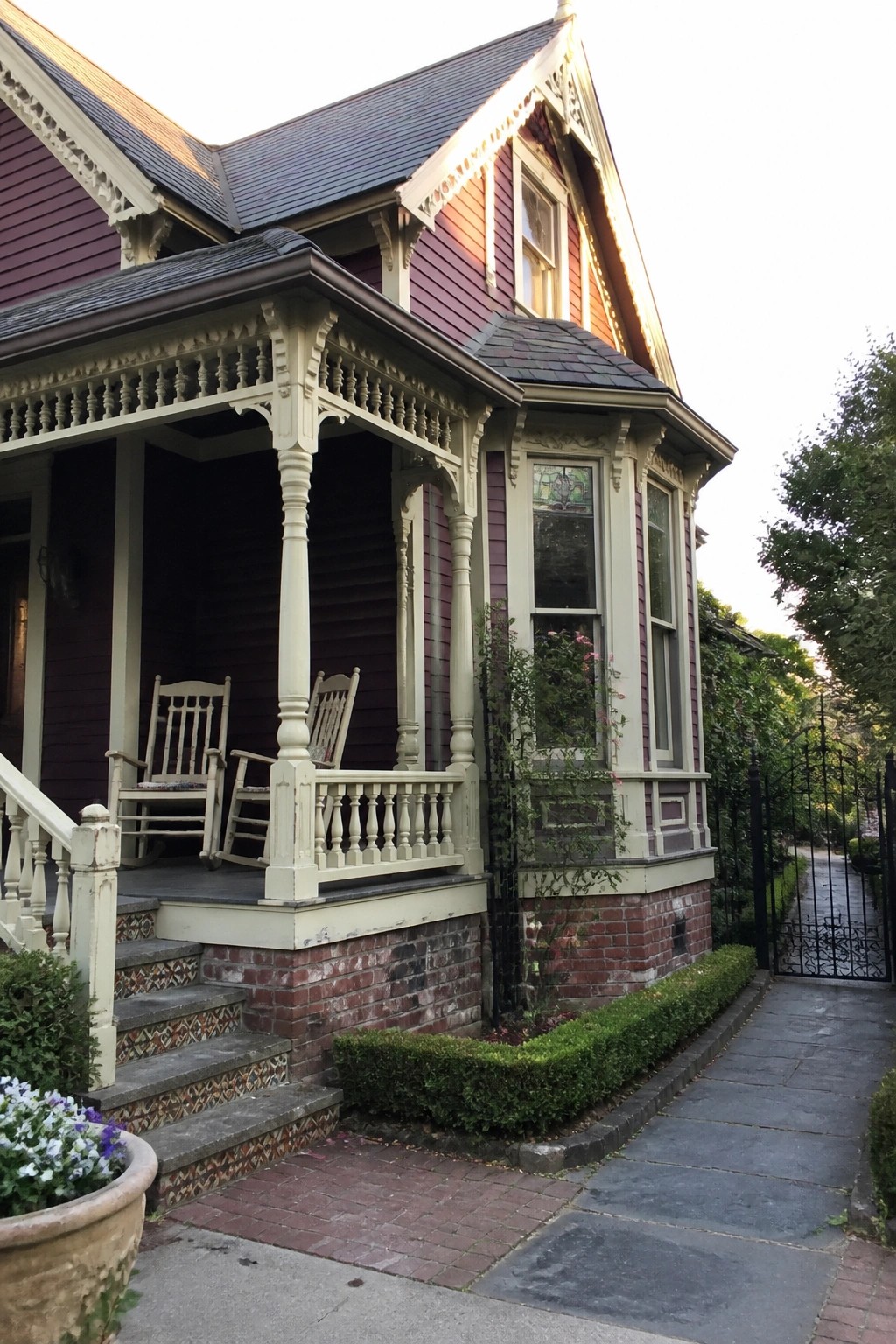 Victorian house with purple siding, ornate porch, rocking chairs, brick steps, and path.
