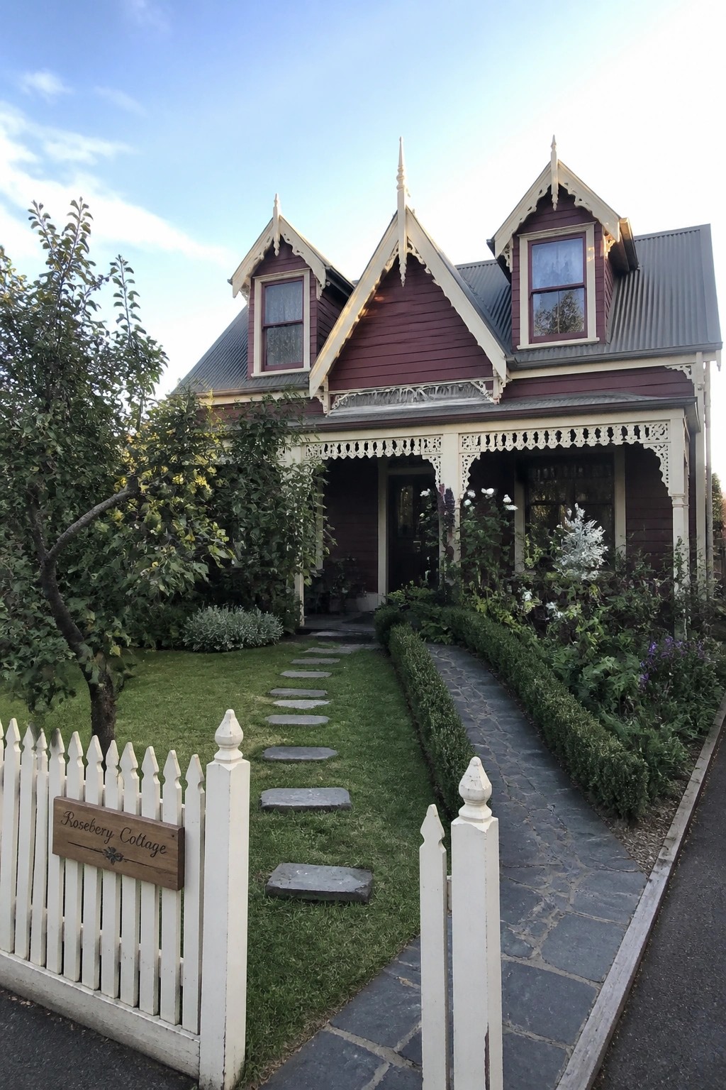 Ornate maroon Victorian cottage with white trim, picket fence, sign, and garden path