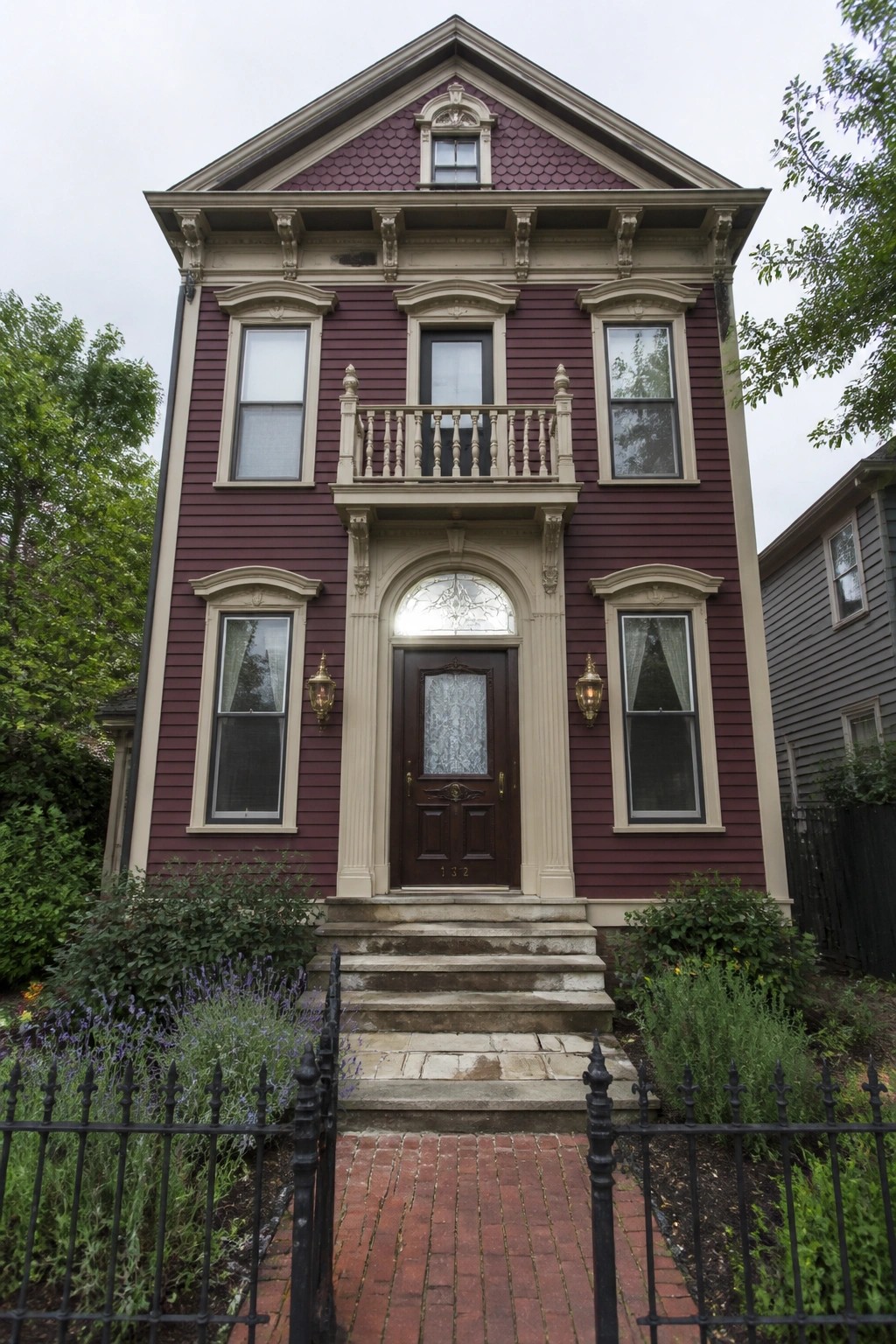 Front view of a three-story burgundy Victorian house with ornate cream trim around windows and door, a small second-floor balcony, and stone steps leading to the entrance.