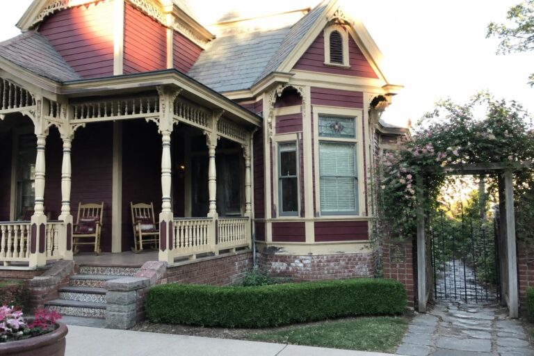 19 Classic Burgundy Victorian House Exteriors Featuring Ornate Cream Trim Victorian house with purple siding, ornate porch, rocking chairs, brick steps, and path.