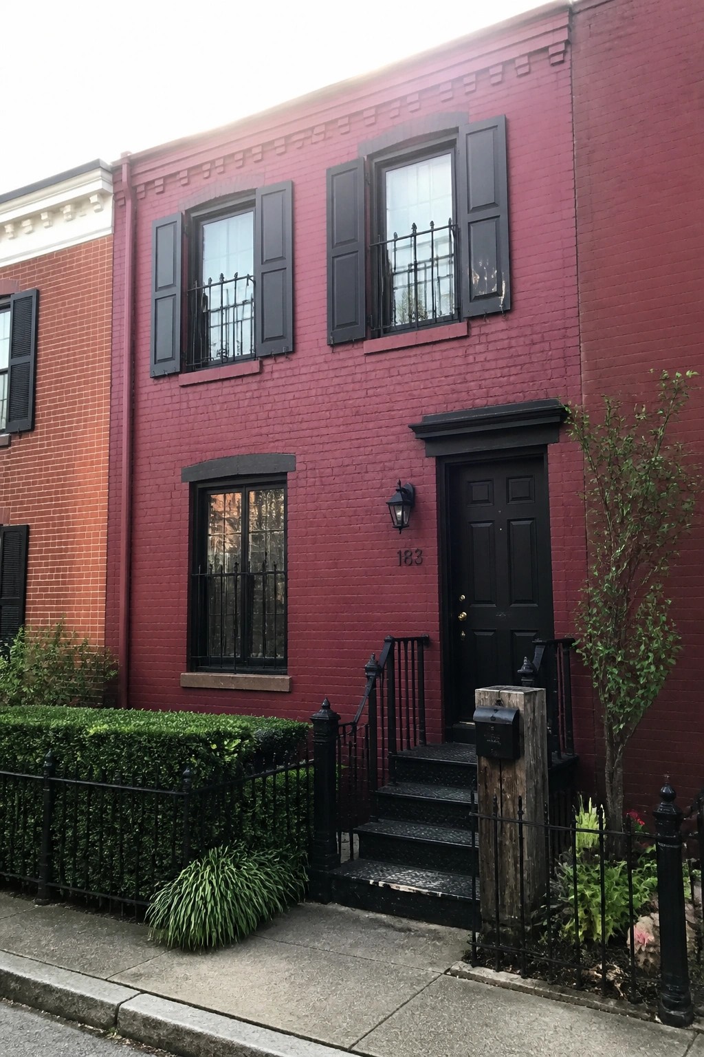 A narrow two-story red brick row house with black shutters, a black front door, and black metal railings at the entrance.