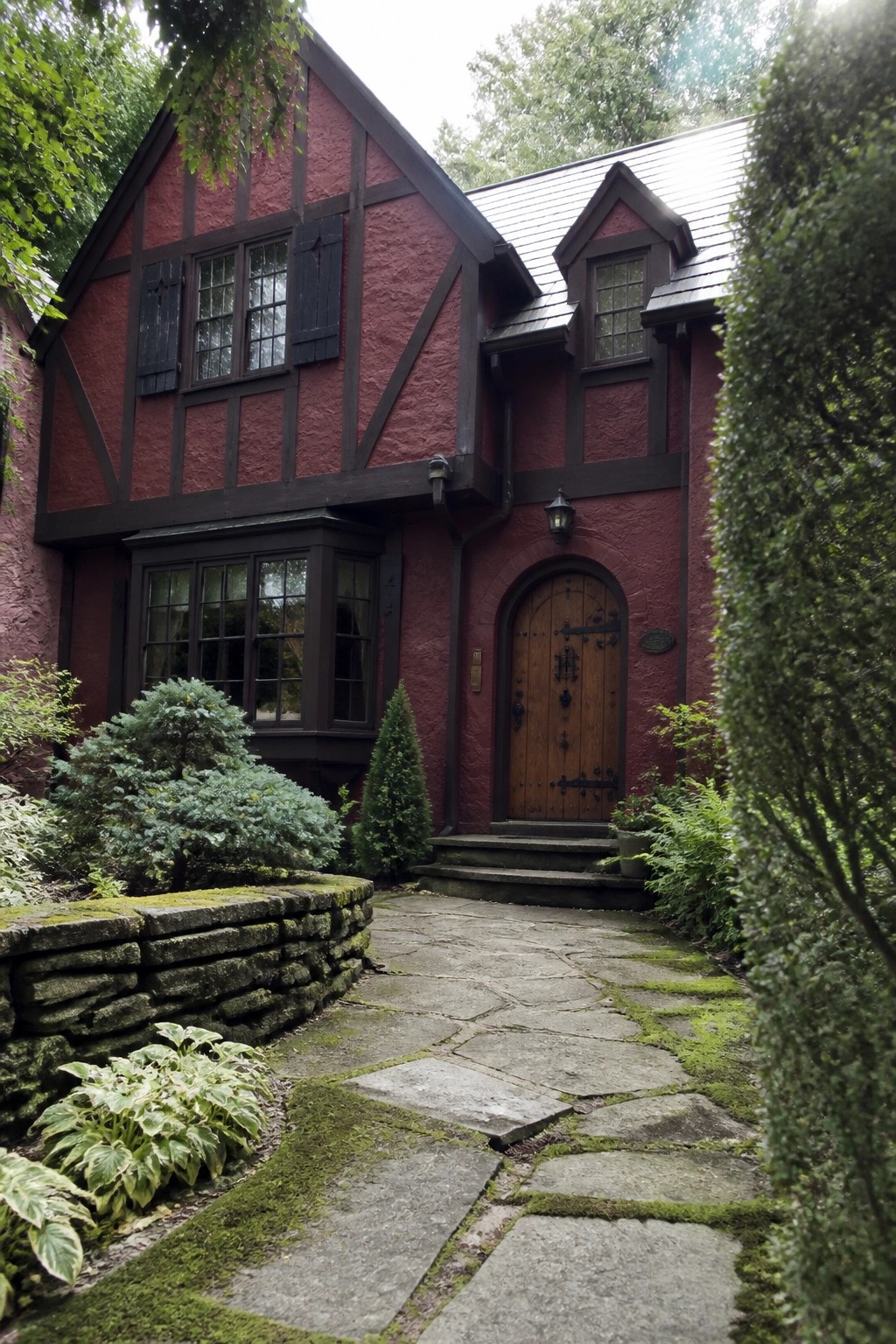 A two-story Tudor-style house with deep red stucco walls, dark wood half-timbering, and black shutters beside a stone pathway and arched wooden door.