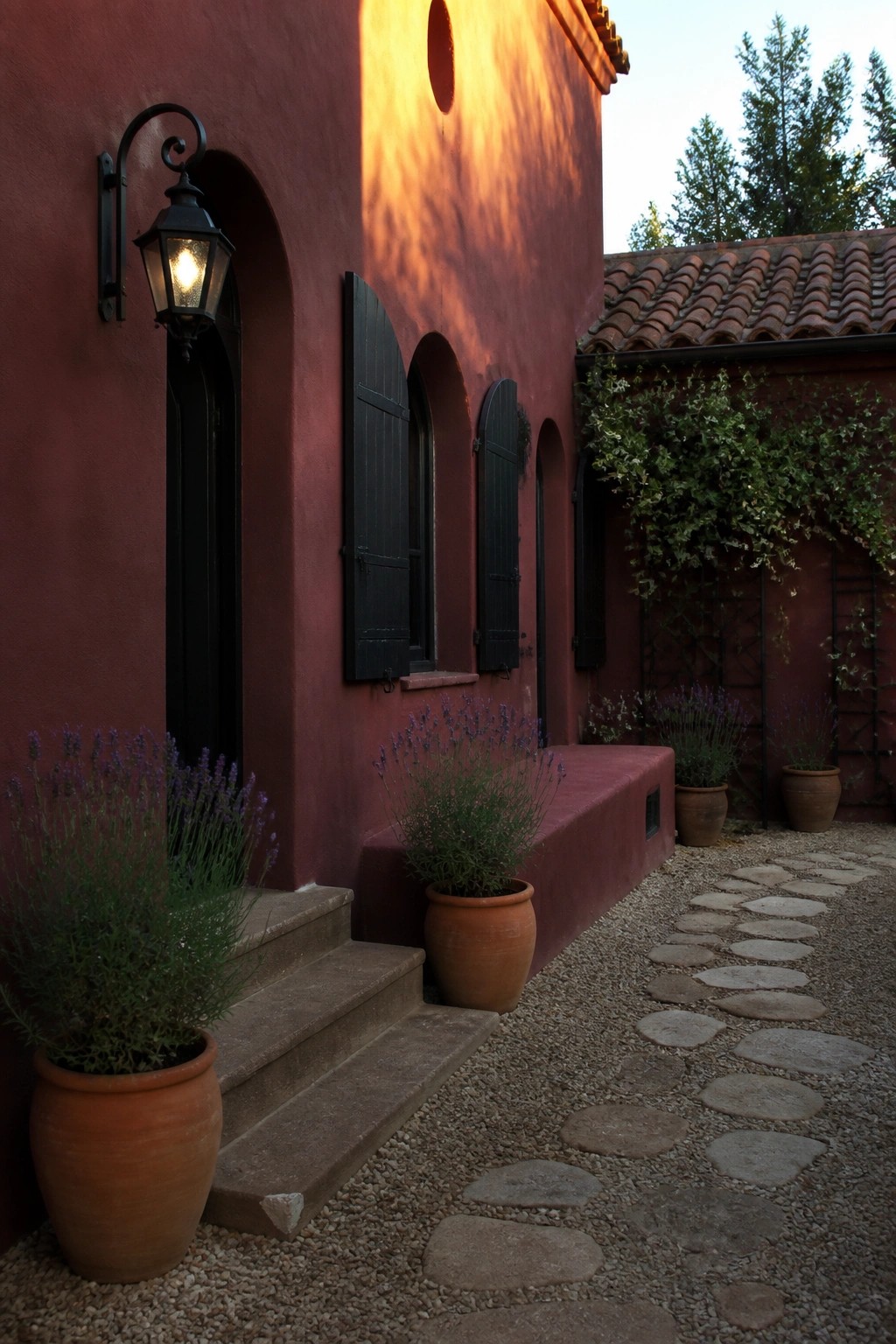 A deep red stucco house exterior with black shutters on arched windows, a lit black lantern beside a dark door, terracotta pots with lavender, and a gravel path with stone stepping stones.