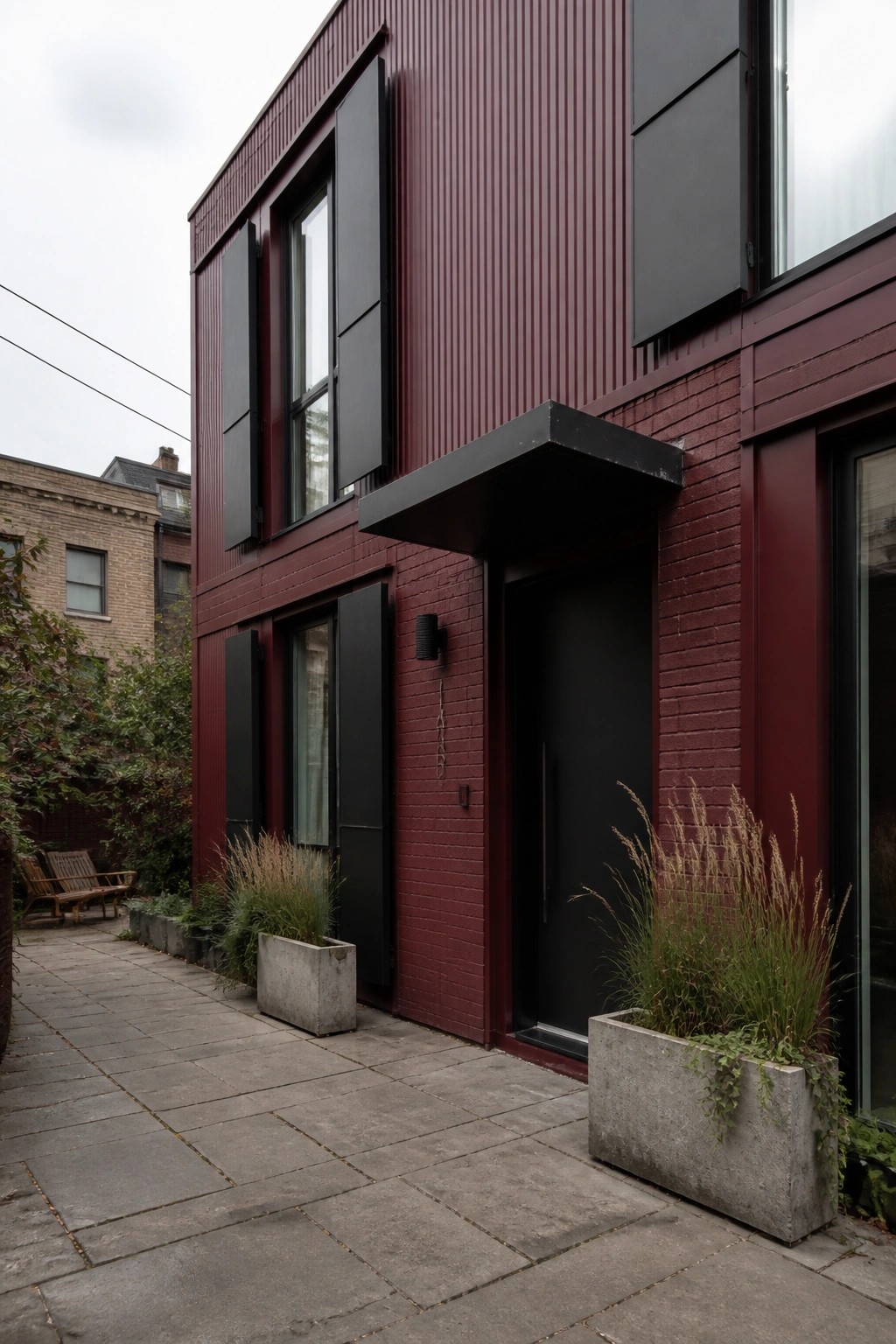 A modern two-story house exterior in deep burgundy with black shutters, a black door, brick and metal siding, and concrete planters along a stone path.