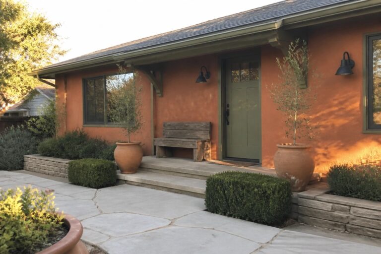 A single-story stucco house with orange walls, an olive green front door and window trim, concrete steps, and terracotta pots holding shrubs next to the entry.