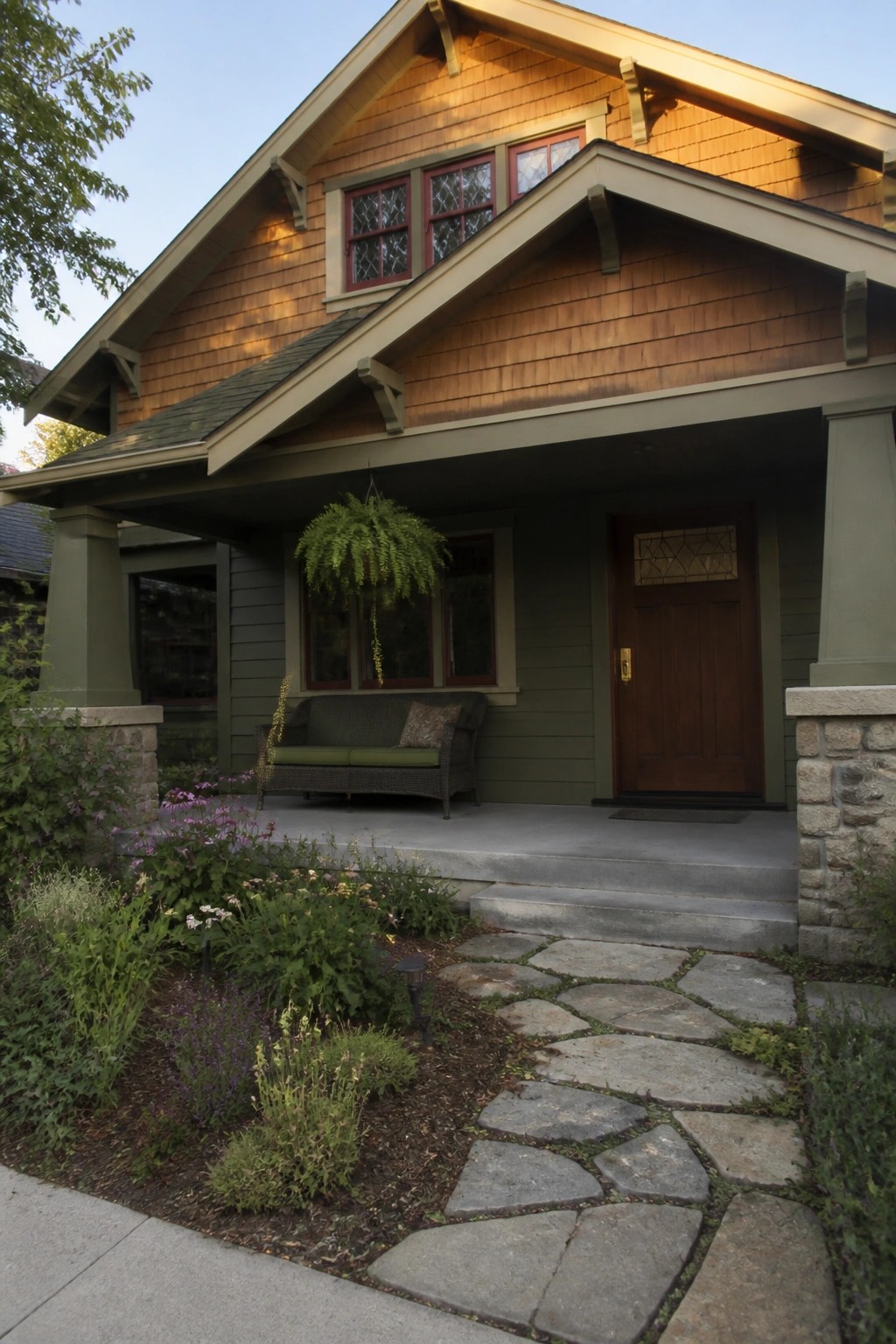 A Craftsman-style house exterior featuring warm brown wood shingle siding, olive green trim and lower siding, a wooden front door, a covered porch with a bench, and a stone pathway leading to the steps.