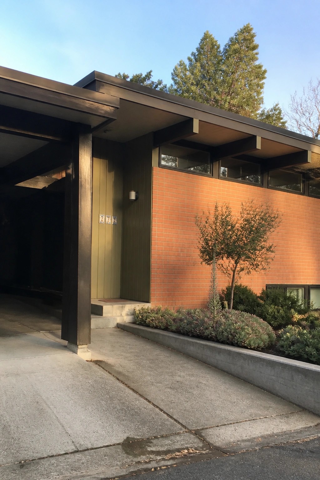House exterior with orange brick wall, olive green vertical siding at the entry, and dark structural overhangs.