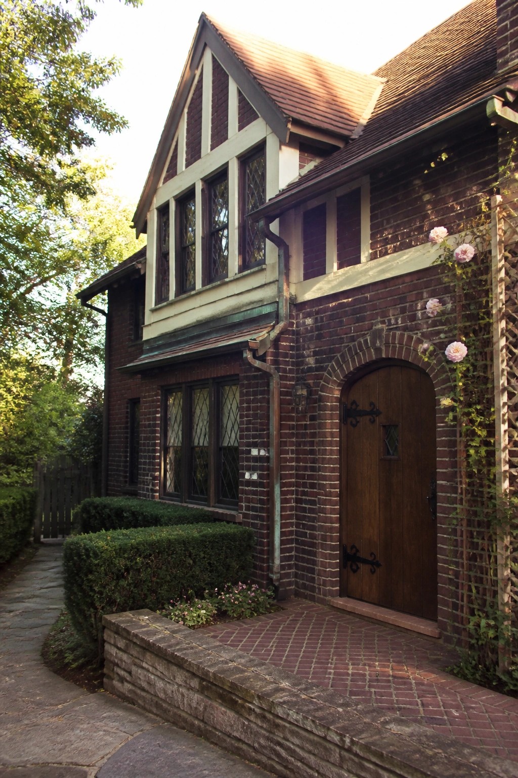 A two-story burgundy brick house with white trim on the gables and windows, a wooden arched front door, and climbing roses along the right side.