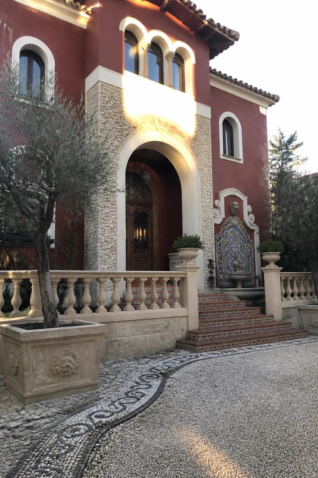 Red stucco villa with arched wooden door, tiled fountain, and gravel courtyard.