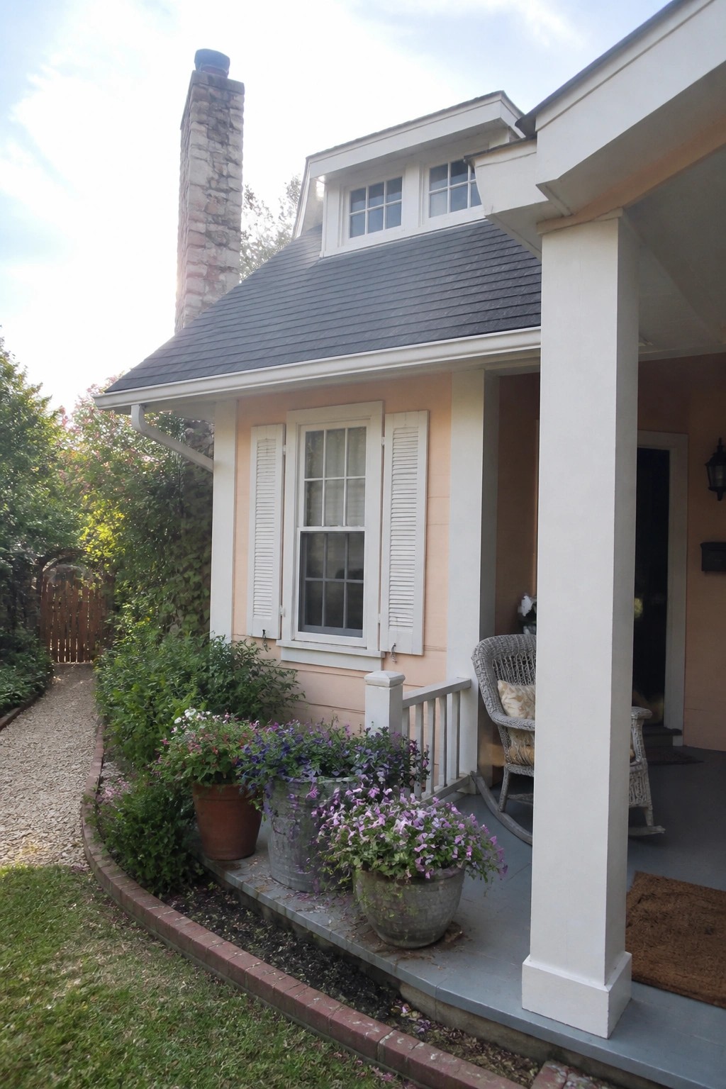 A peach-colored house exterior with white shutters, a dark roof, a stone chimney, and potted flowers along the front porch.