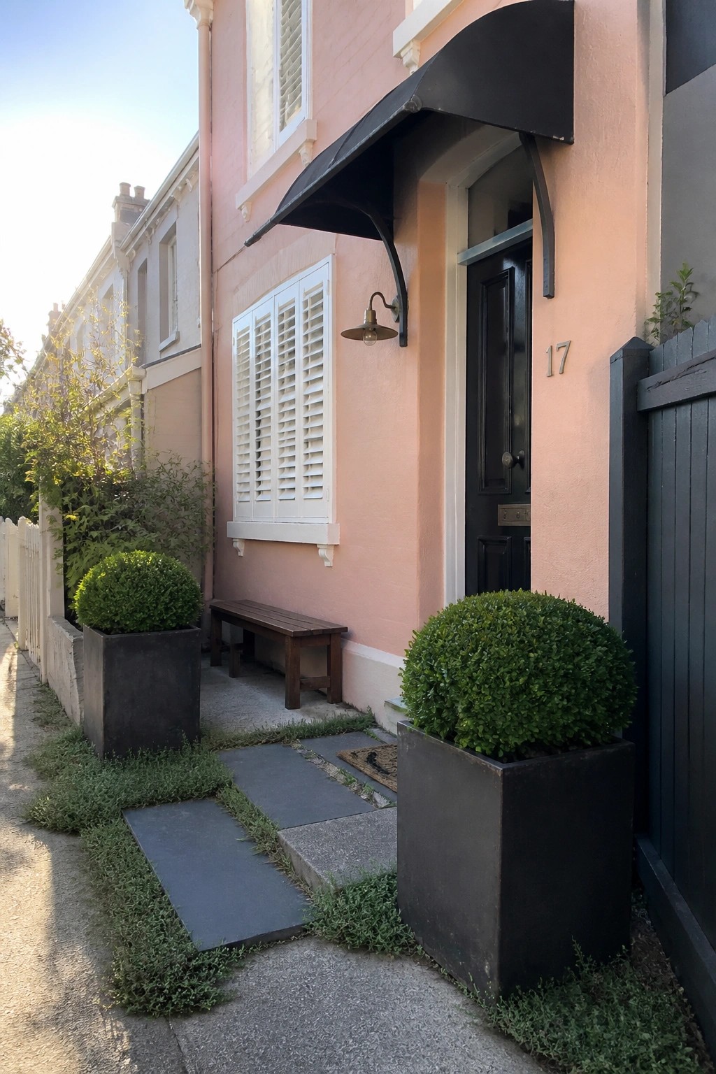 Pink terraced house with black door 17, awning, white shutters and potted shrubs.