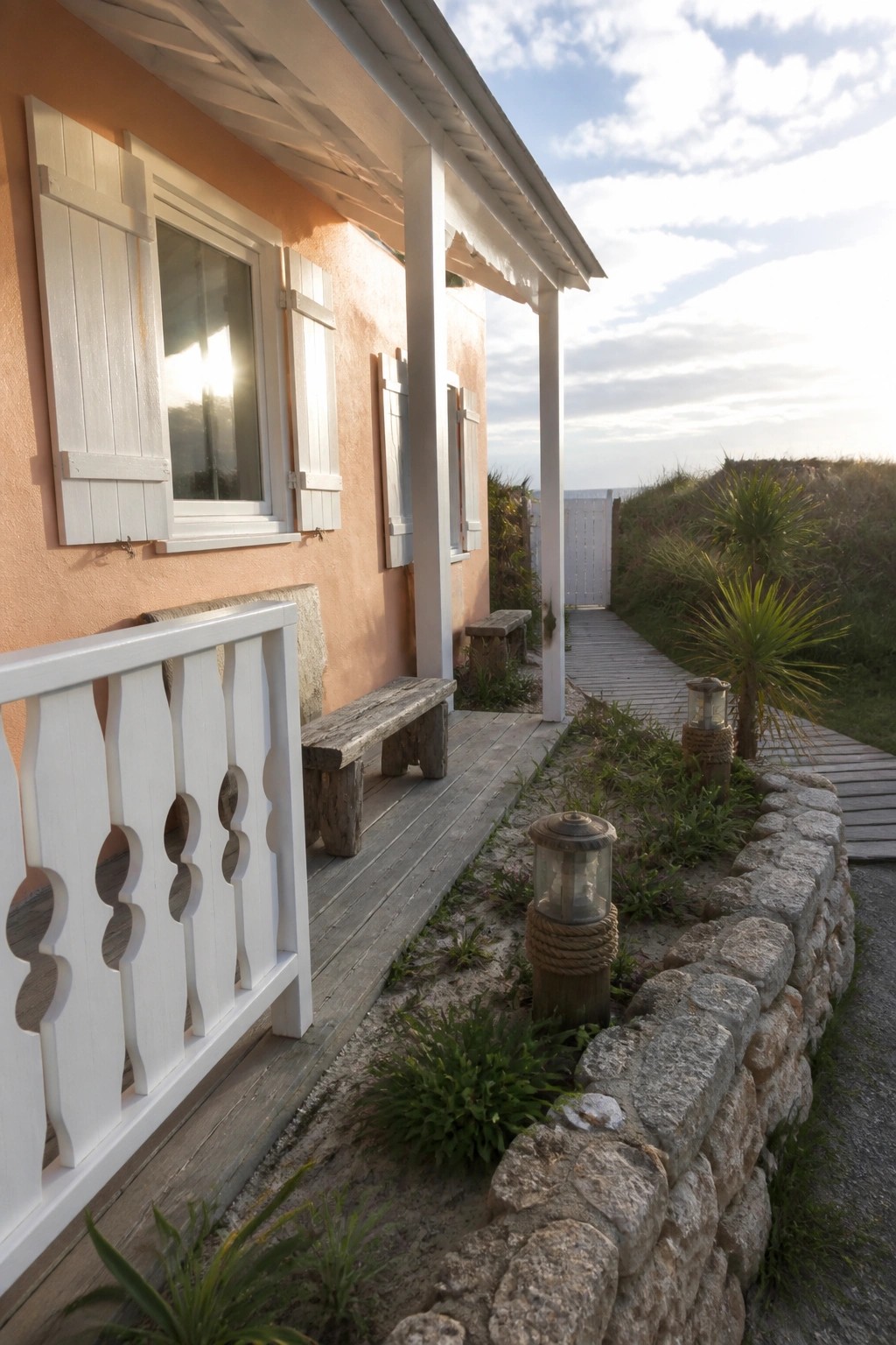 Peach beach house porch with white shutters, wooden bench, stone lanterns, and boardwalk.