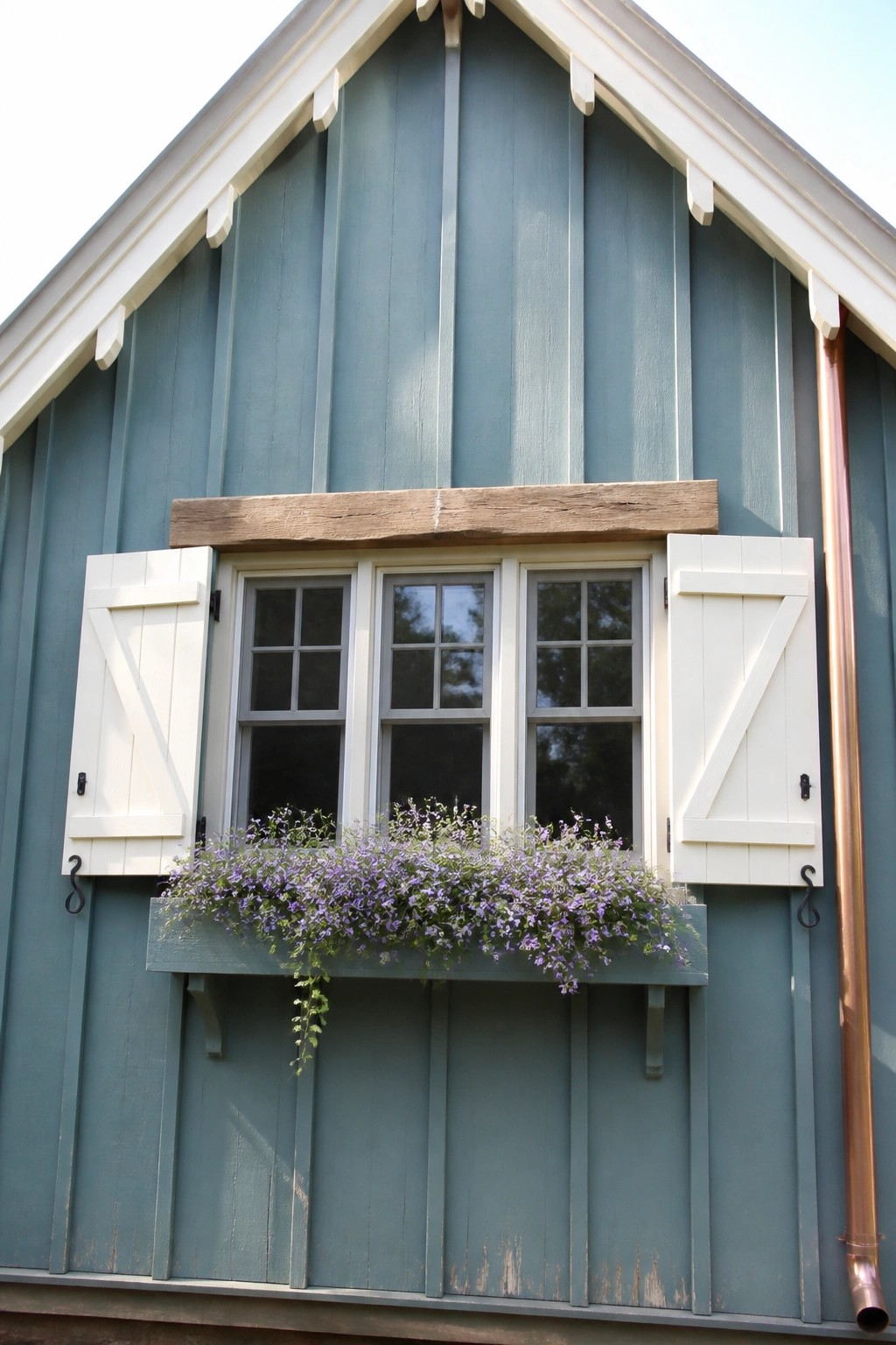 Teal wooden house exterior with white shutters open on a three-section window and a flower box filled with purple blooms below.