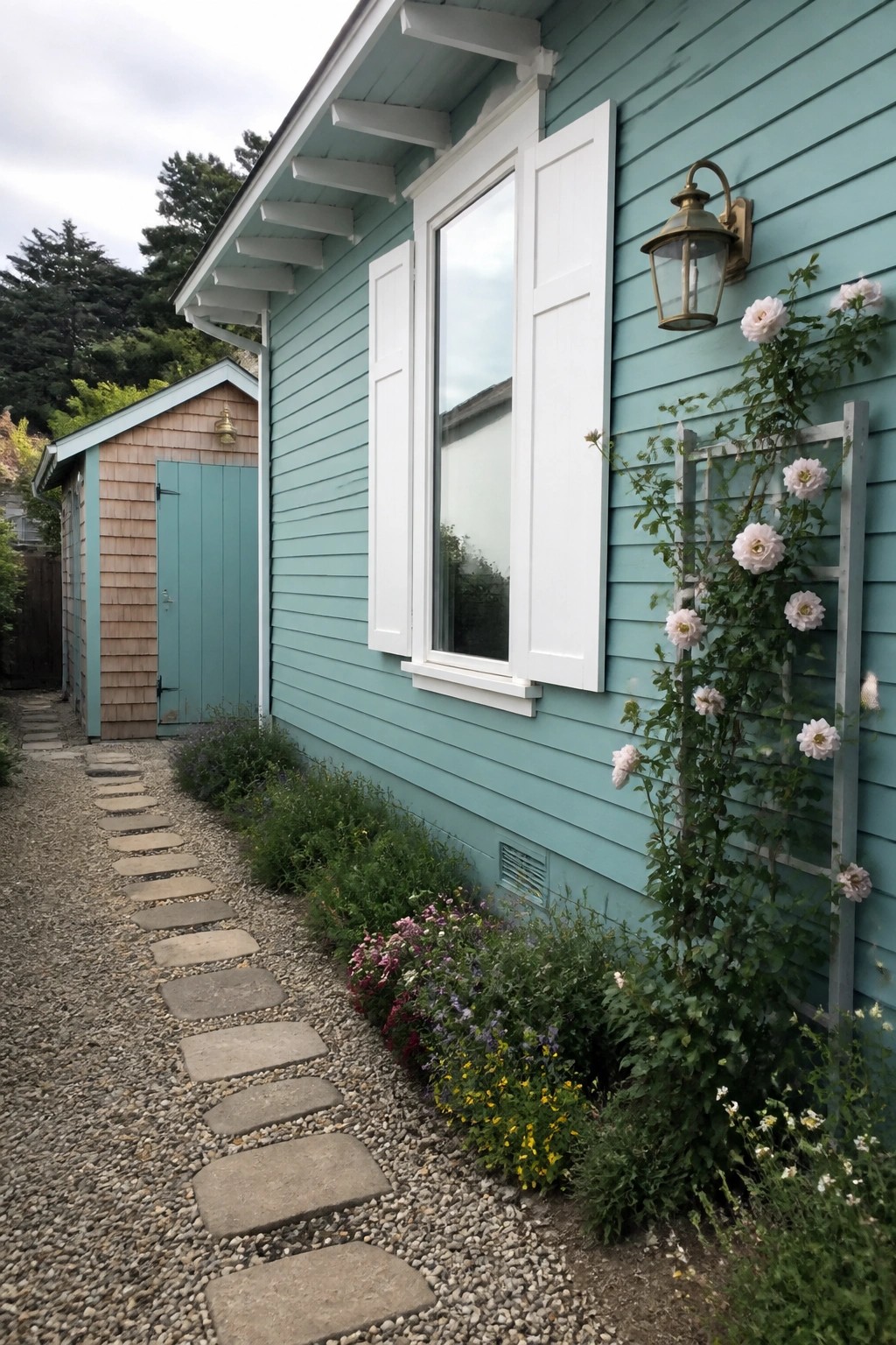 A light teal house exterior with white shutters on a window, a gravel path lined with stepping stones, and white roses on a trellis.