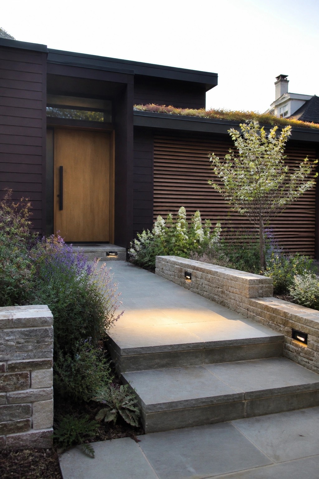 Modern home entrance with wooden door, stone steps, and lush garden landscaping.