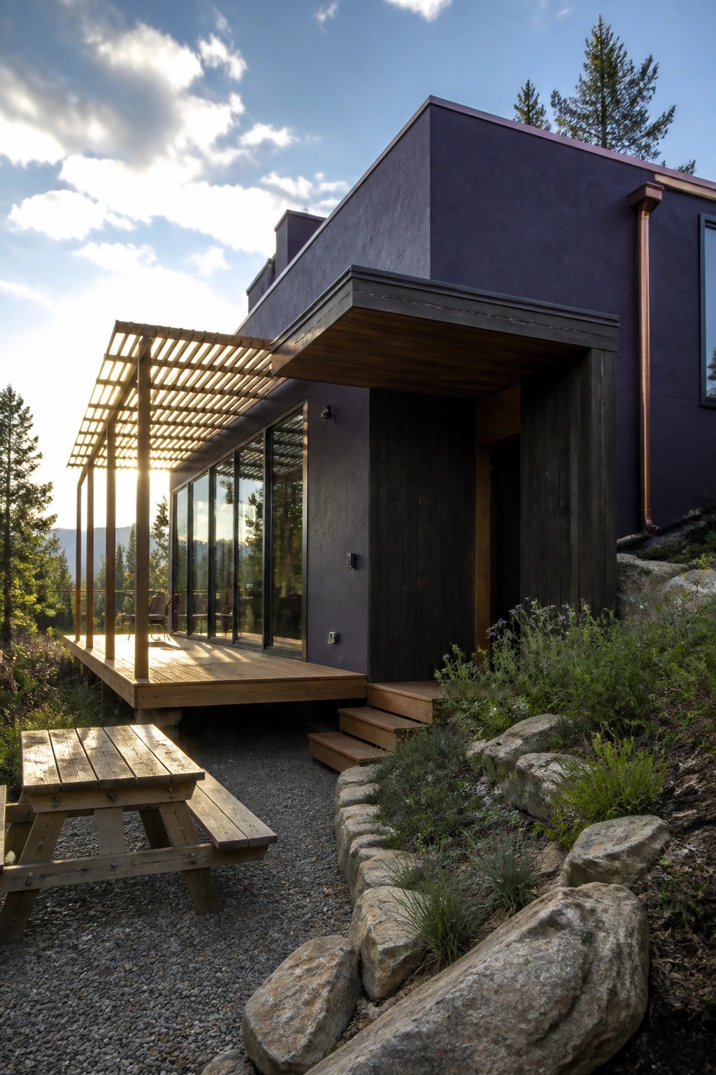 A modern house with deep purple exterior walls, a wooden deck and pergola, large glass windows, and stone landscaping on a hillside.