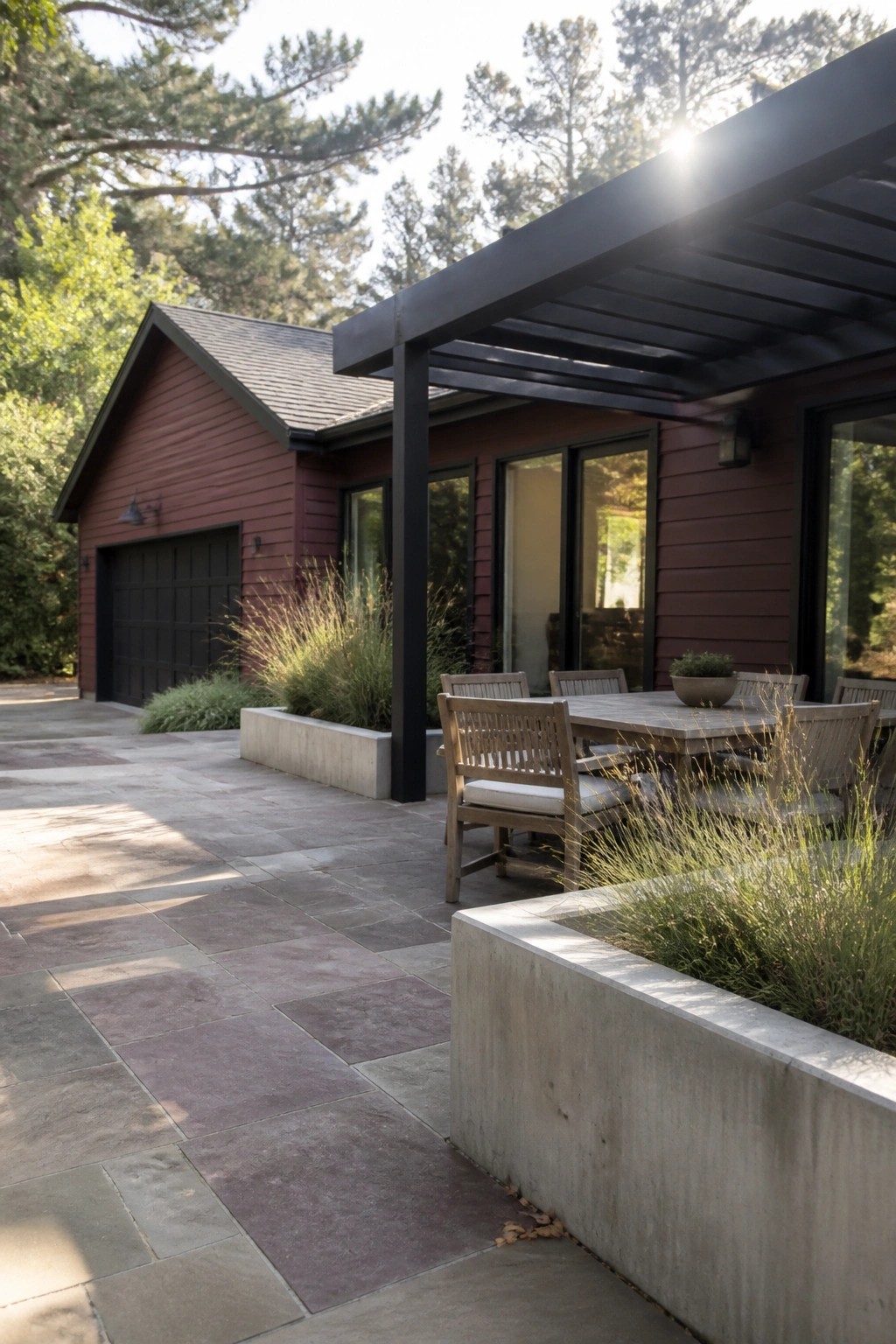 A maroon house with black trim shows a stone patio dining area under a black pergola with concrete planters and surrounding grasses.