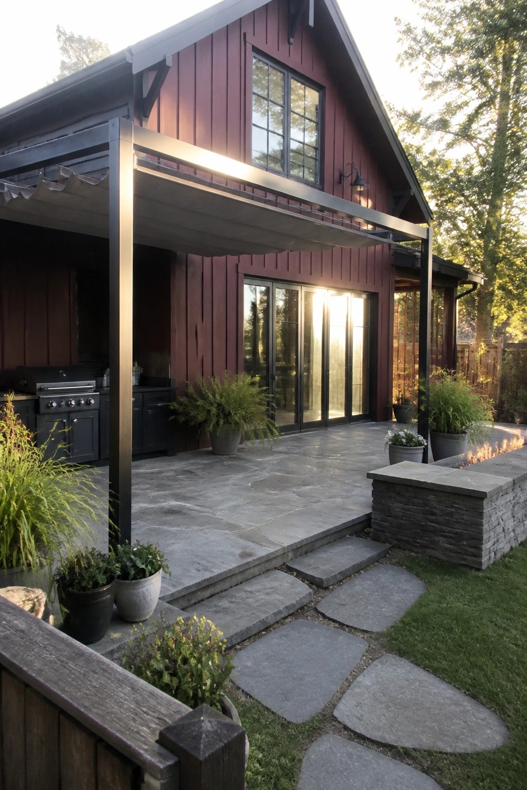 A maroon house with matte black trim features a covered stone patio with an outdoor kitchen, glass doors, potted plants, and a stone fire pit, connected by a flagstone path.
