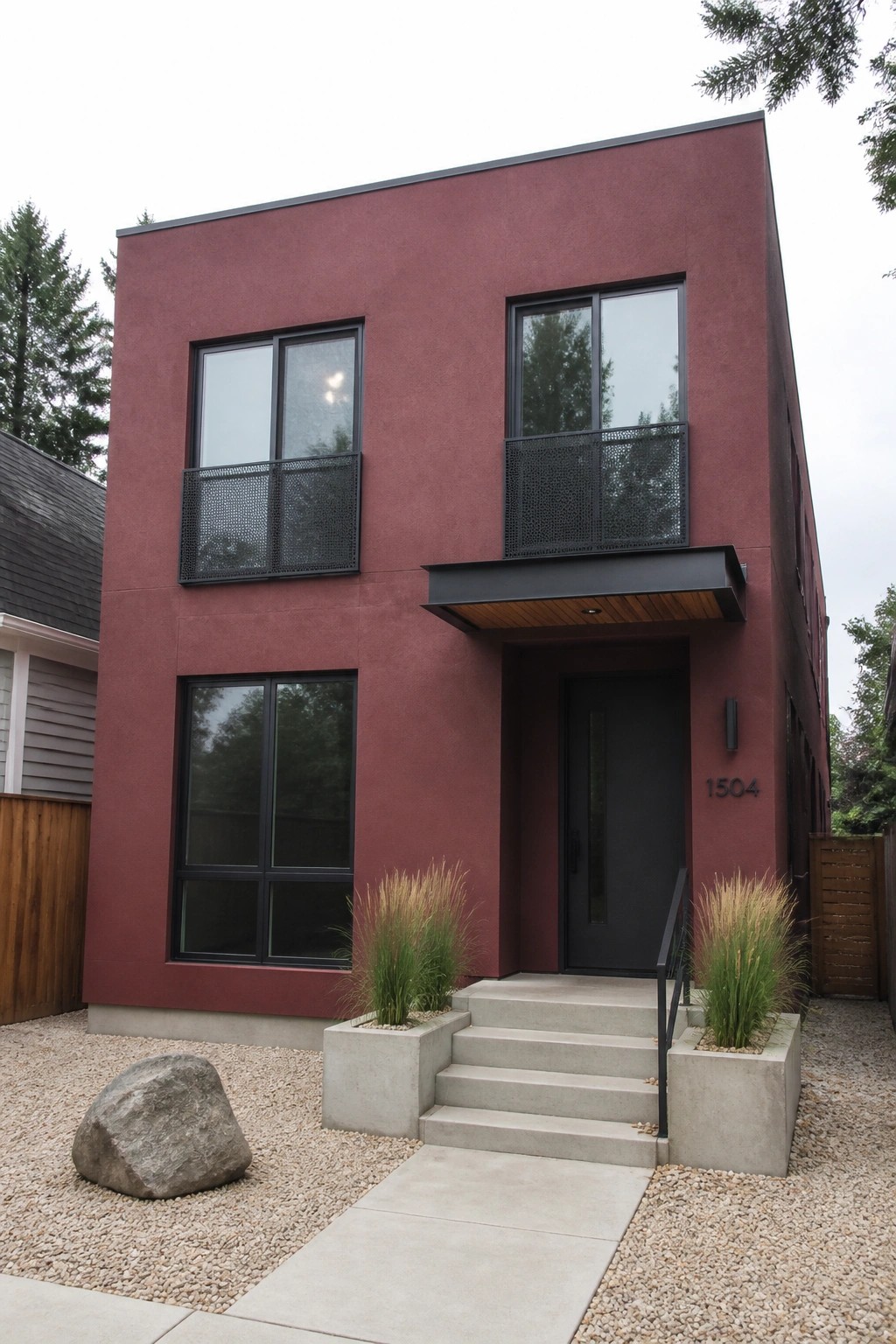 A two-story modern house with a deep maroon stucco exterior, matte black window frames, a black front door under a small canopy, and concrete steps flanked by concrete planters.