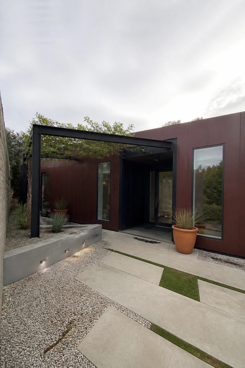 Modern house exterior with maroon siding, matte black window and door trim, black metal entry structure, and concrete pavers with gravel and potted plants.