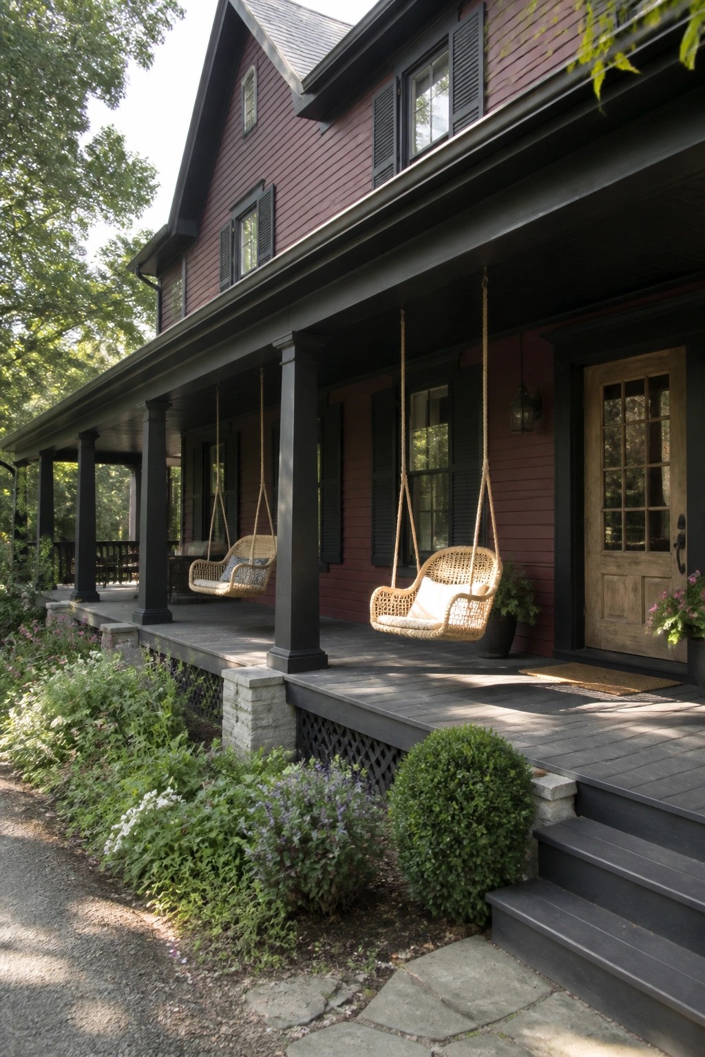 Red house with black trim, two hanging wicker swings on shaded porch with garden.