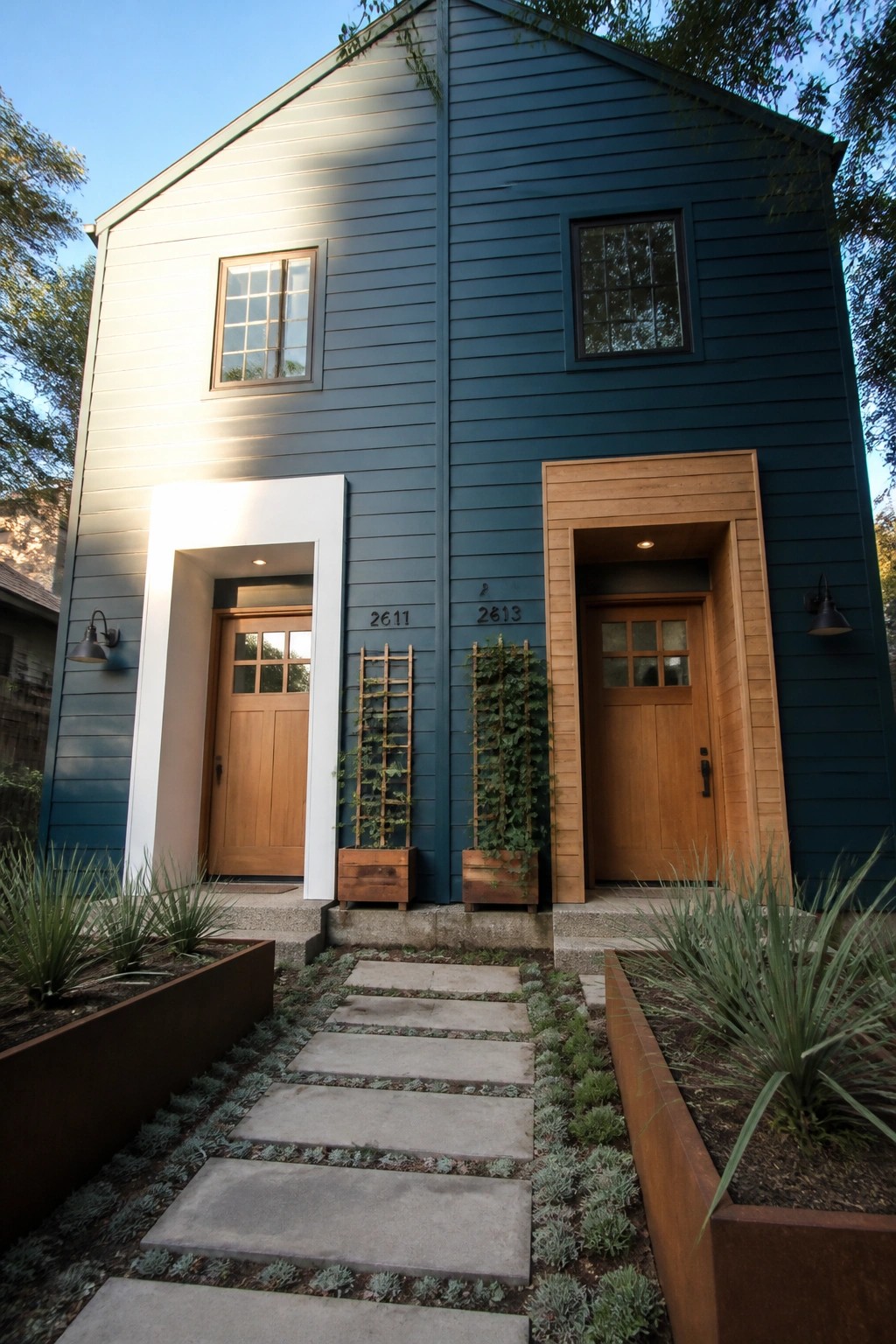 Front view of a two-story house with deep teal horizontal lap siding, two wooden doors with different surrounds, and a concrete path leading to the entrances.