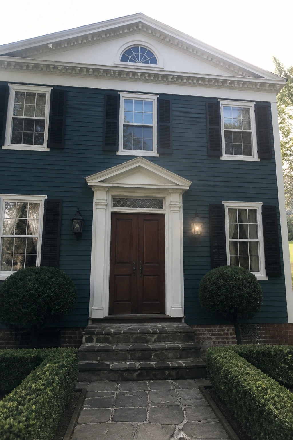 Front view of a two-story house with deep teal lap siding, white trim, black shutters, and a white pediment over the double front doors.