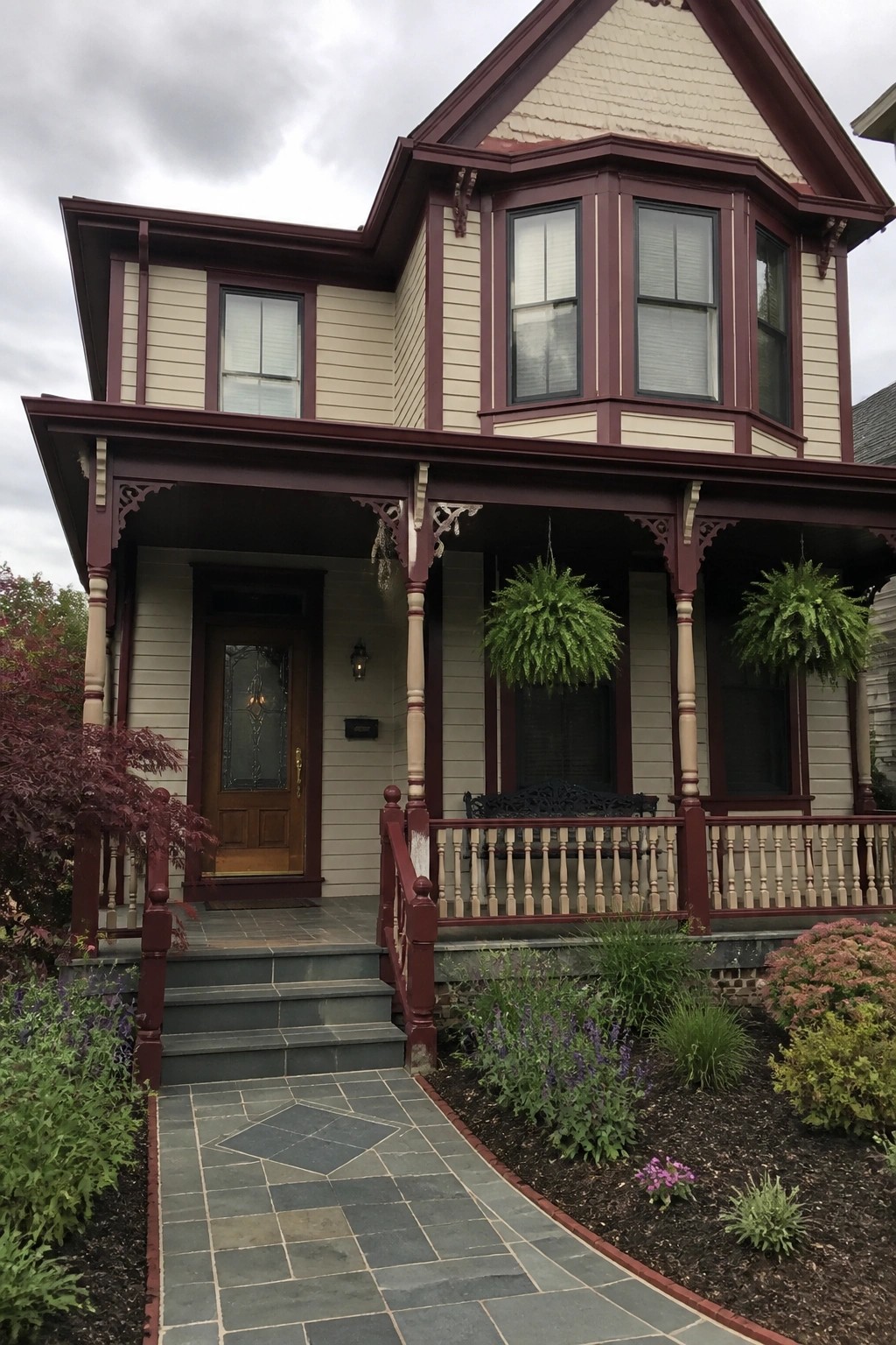 A two-story house exterior with beige siding, deep burgundy trim, a wooden front door, and a covered porch featuring turned wood posts and railings.