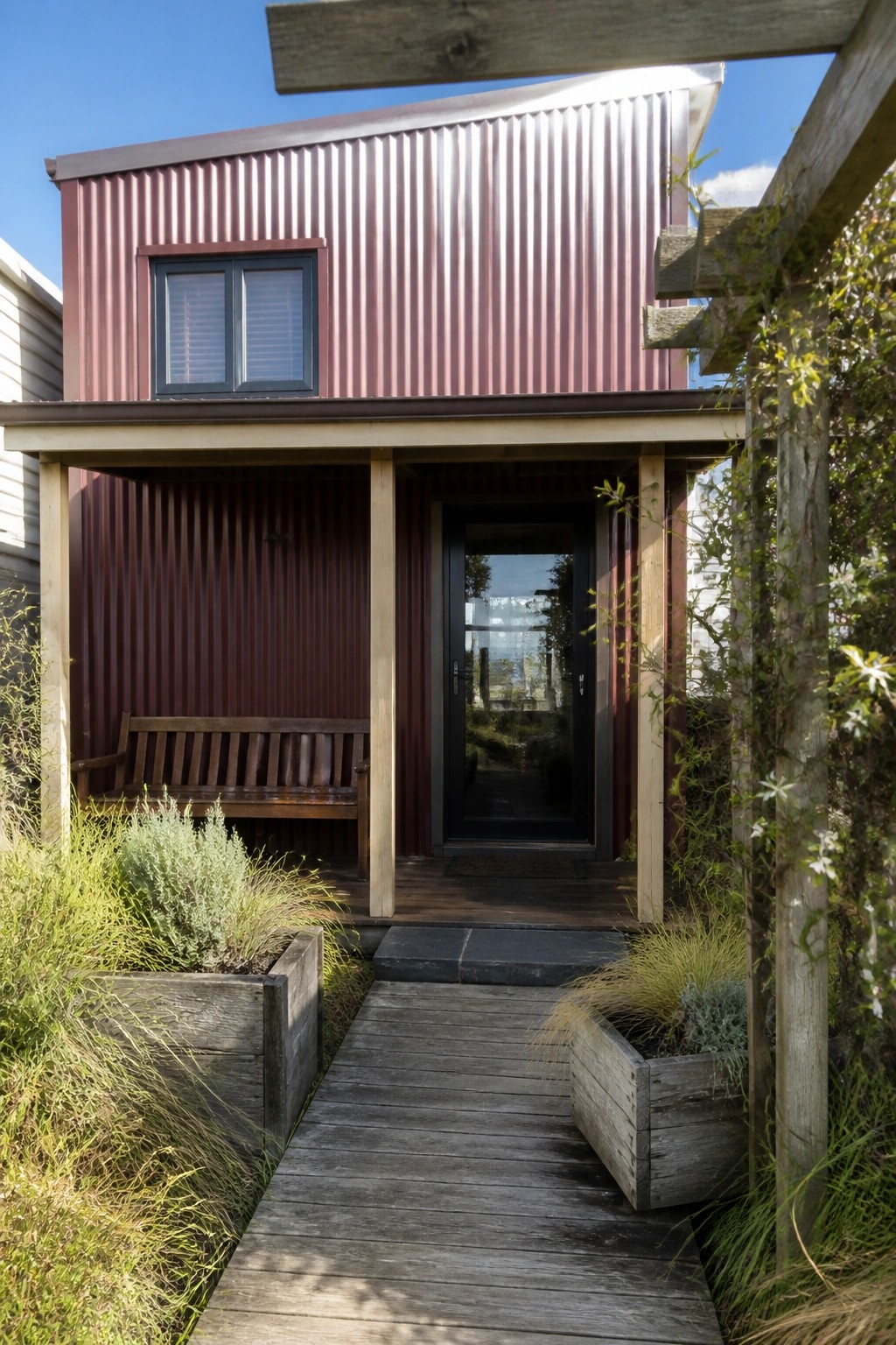 A burgundy corrugated metal house with a timber porch, wooden bench, deck path, and planters beside the entry door.