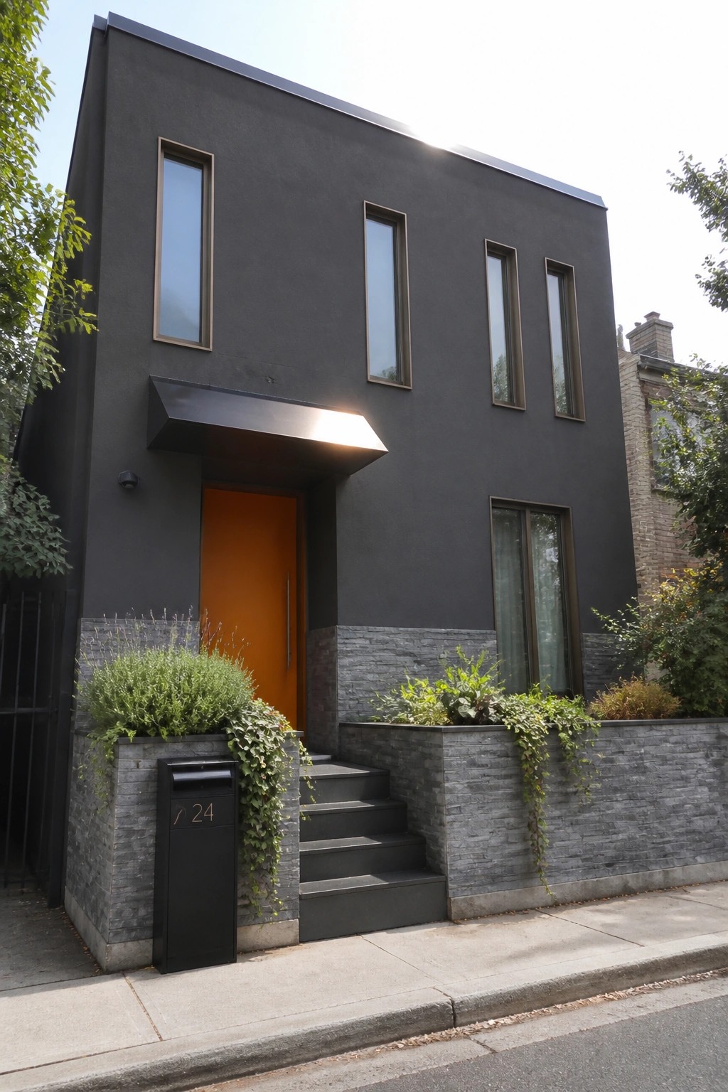 A modern two-story house with dark gray exterior walls, an orange front door under a black overhang, narrow vertical windows, and gray stone cladding at the base.