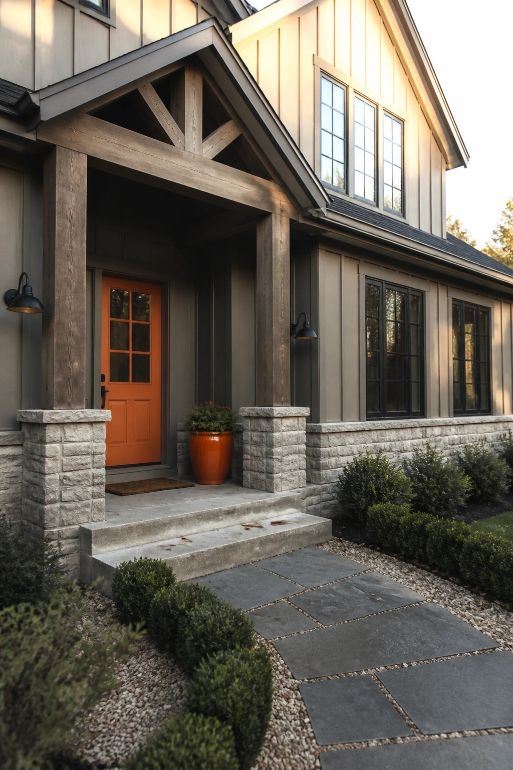 Front exterior of a gray house with an orange door, wooden beams, stone pillars, and a stone pathway leading to concrete steps.